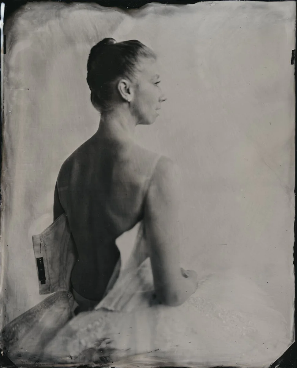 Black-and-white portrait of a woman with short hair, sitting with her back to the camera and her face in profile, with a blank light background.