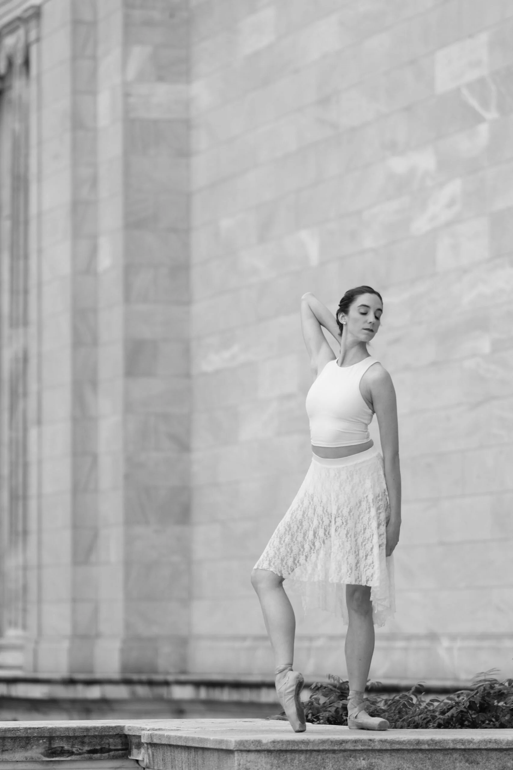 A black and white photo of a ballerina in a dance studio, standing in a pose with arms raised and head tilted slightly, wearing a sleeveless top and a long skirt, with ballet shoes on her feet. The background features a large brick wall.