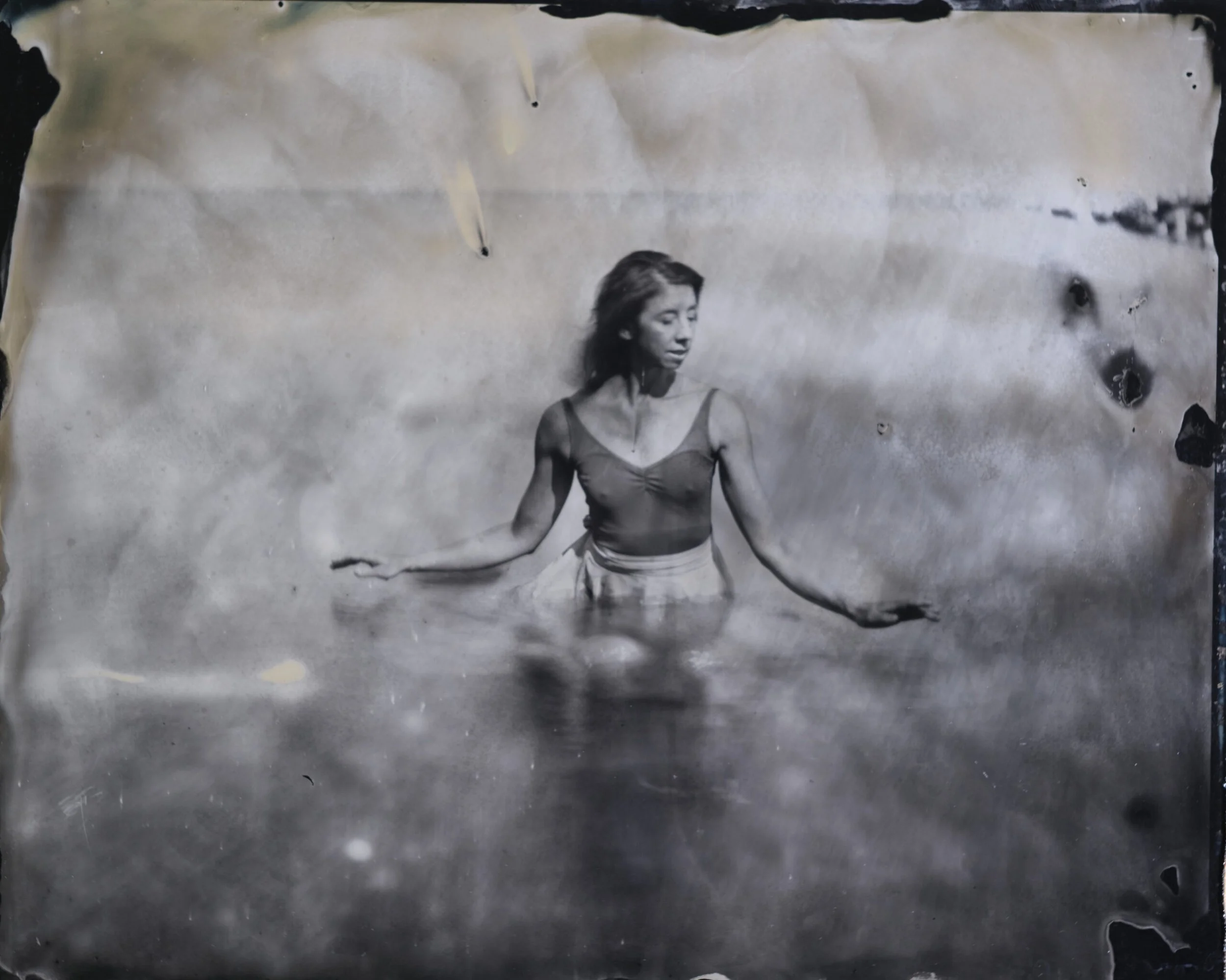 A black-and-white photograph of a young woman with short hair, wearing a sleeveless top, looking down thoughtfully in front of a plain background. The image is on a distressed sheet of paper with burnt edges.