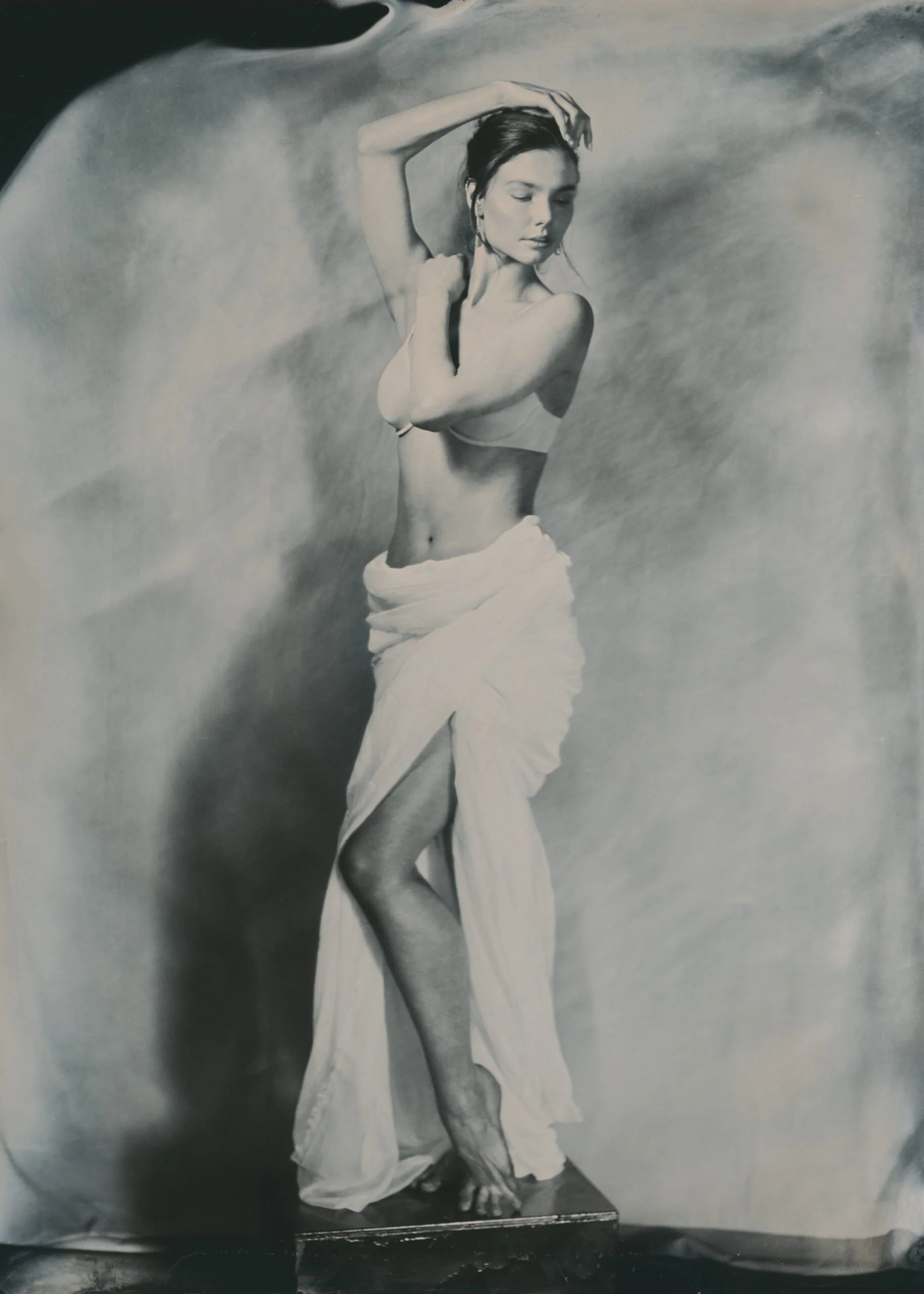 A black and white photo of a woman with closed eyes, standing with her arm raised above her head, wearing a bikini top and a long, flowing skirt, posing against a cloudy background.