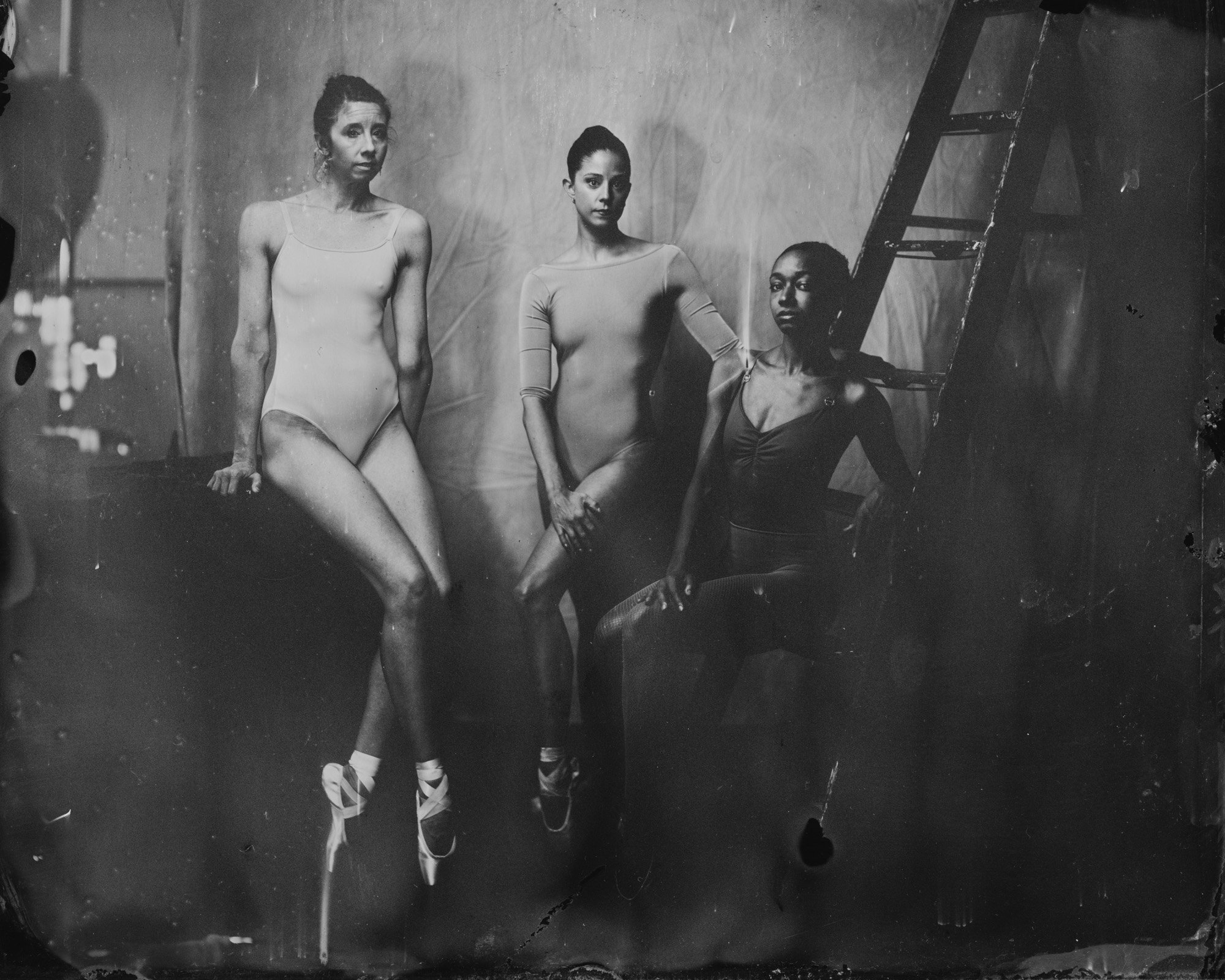 Three ballet dancers in rehearsal, standing in a studio with a plain backdrop. One dancer is sitting on a bench, two are standing nearby. All are dressed in leotards and ballet shoes, preparing for practice.