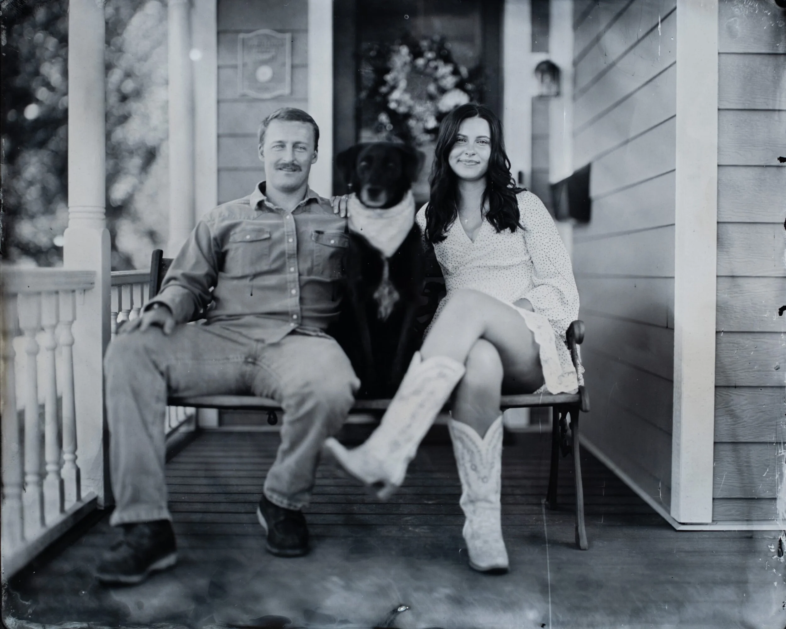A black and white photo of a man, a woman, and a dog sitting on the porch of a house. The man has a mustache and is wearing a button-up shirt, the woman has long dark hair and is wearing a dress, and the dog is a black Labrador sitting between them.
