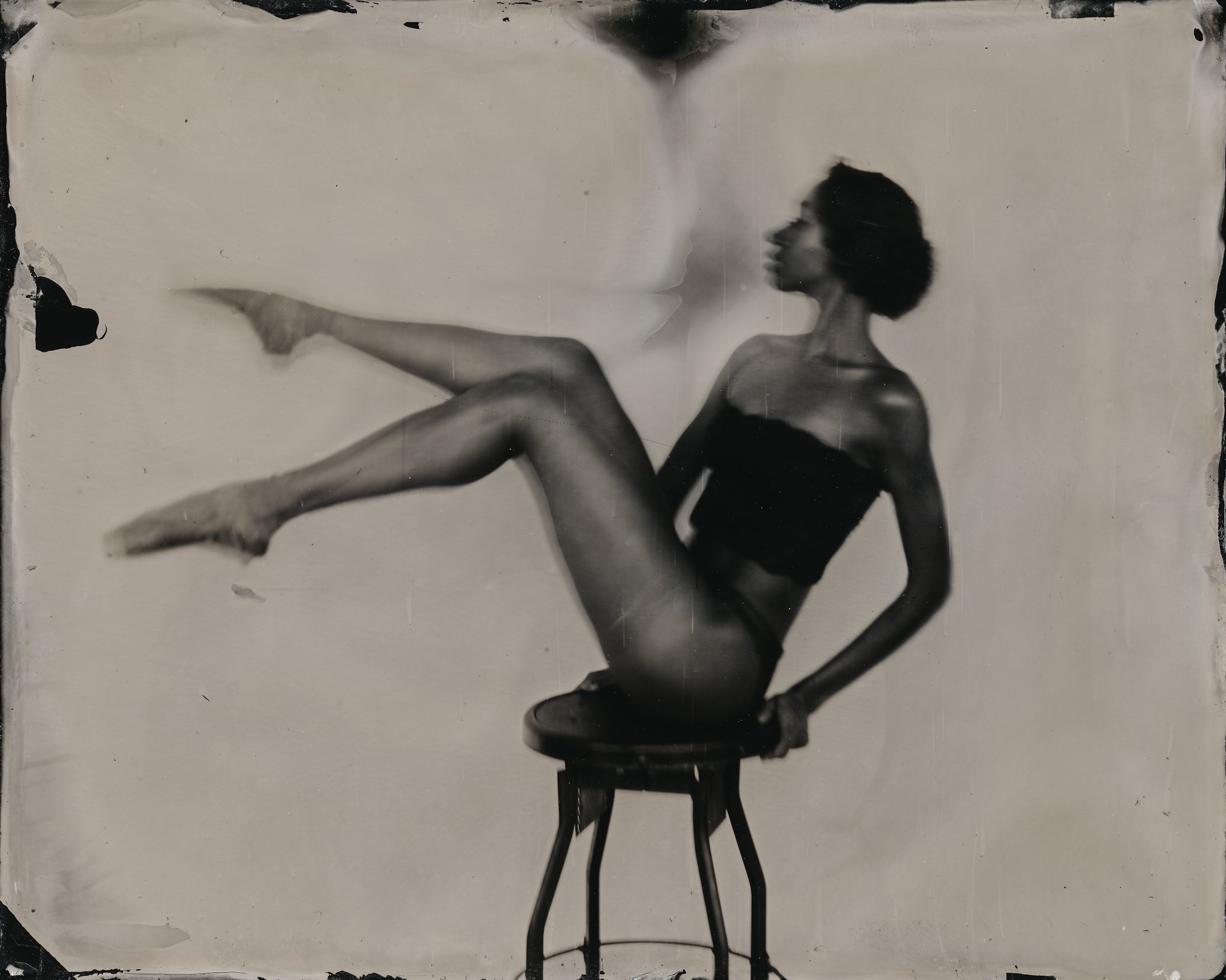 Black woman with curly hair sitting on a chair with her legs extended, pointing her finger, against a plain background.