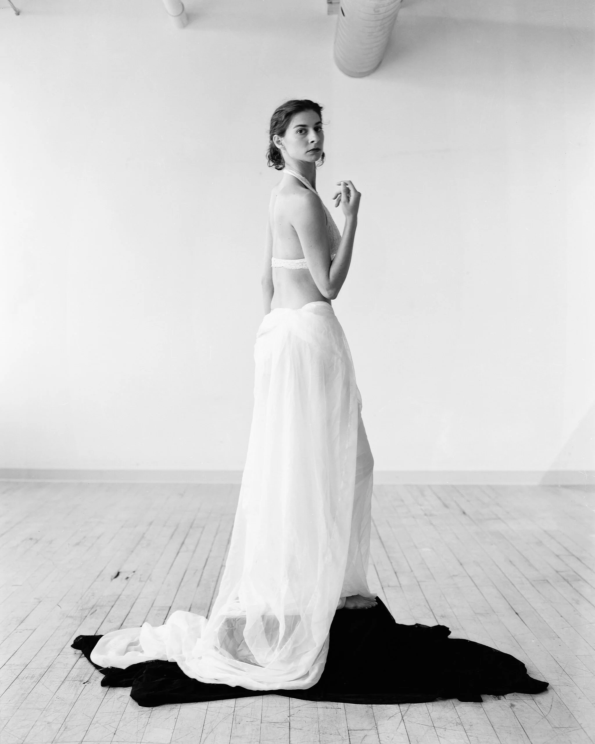 A woman standing on a wooden floor with a plain white wall behind her. She is dressed in a flowing white dress with a strapless bodice and a voluminous skirt, leaning to her right with her left hand near her chest, and looking directly at the camera.