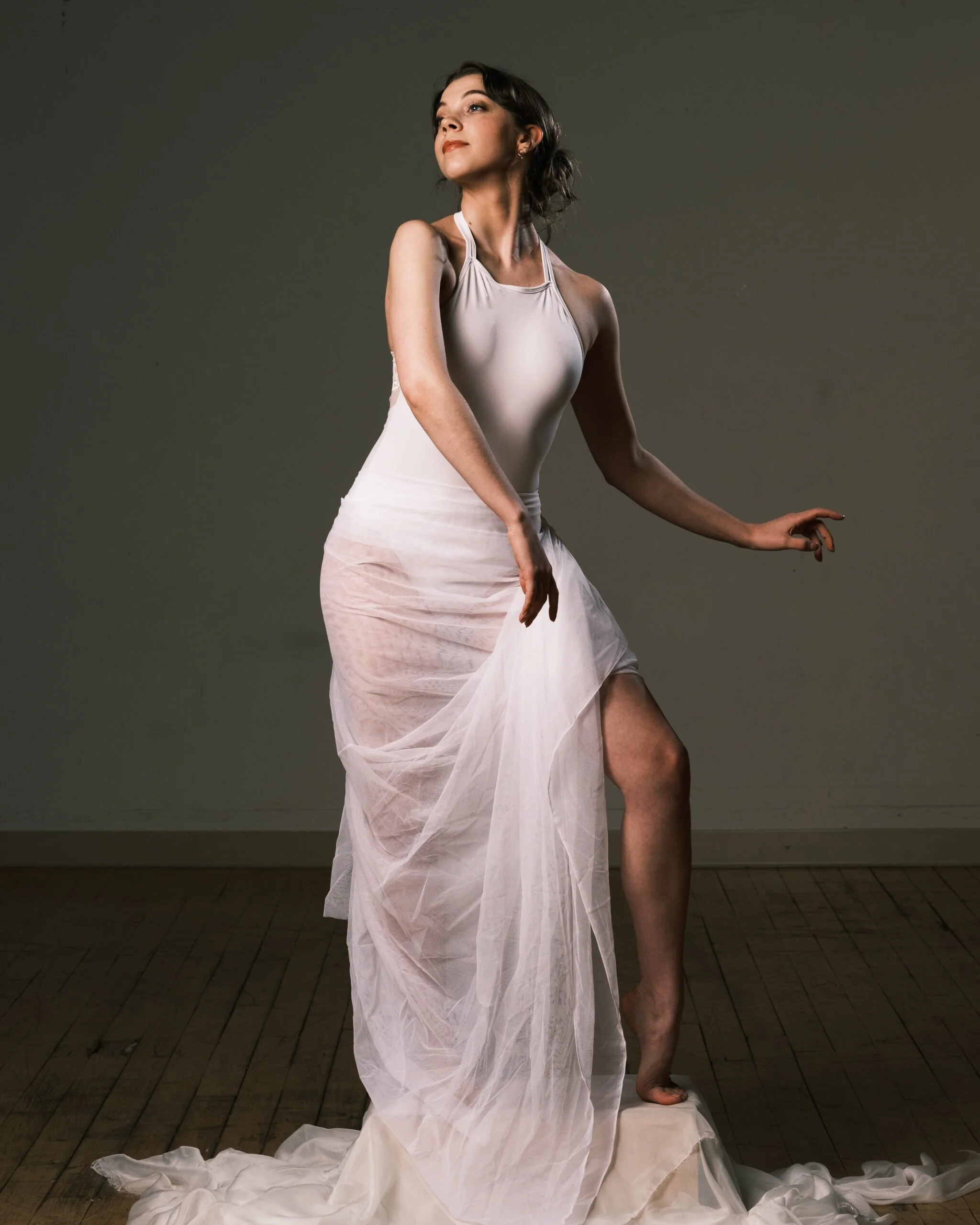 A woman in a white dress stands on a wooden floor against a plain background. She has dark hair, looks to her right with a confident expression, and is partially posing with her right arm slightly bent and her left arm extended downward, her Leg is v