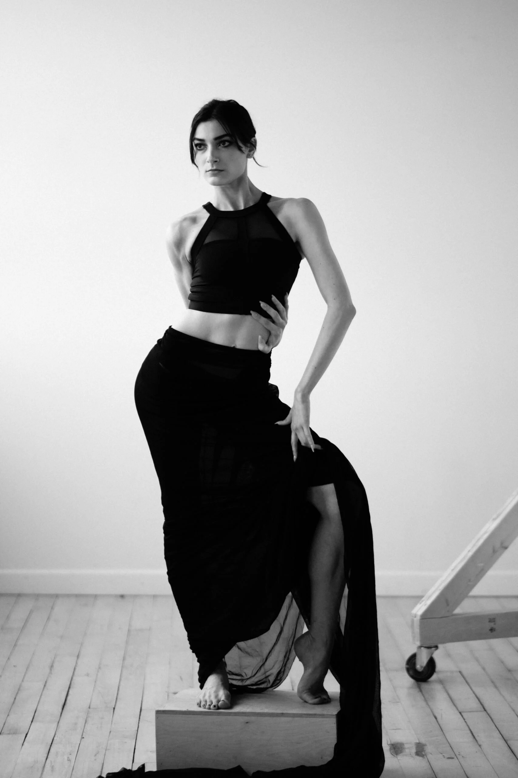 A woman with dark hair and a sleeveless black top, standing on a wooden floor in front of a plain white wall, poses with her right hand on her waist and her left hand resting on a man dressed in dark clothing, kneeling and holding a piece of fabric o