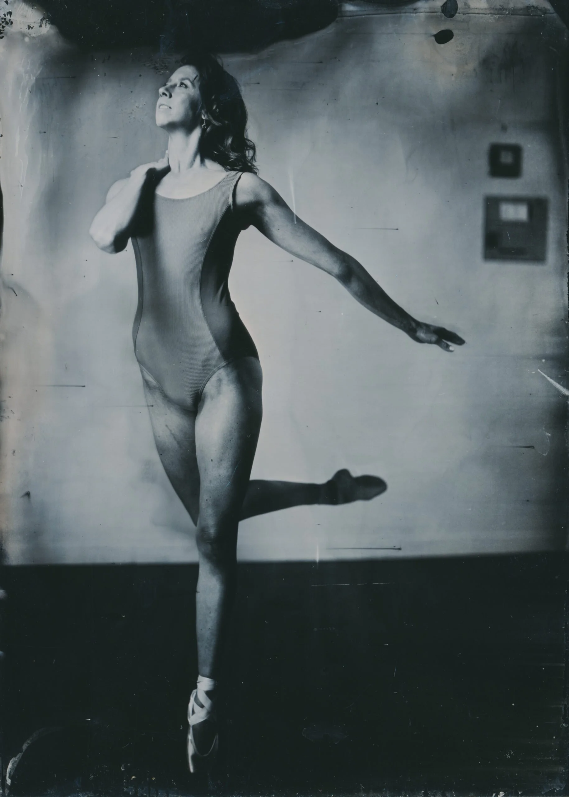 Black and white photograph of a ballet dancer practicing with one foot on the ground and the other extended behind her, wearing a form-fitting leotard and ballet shoes, in a dance studio with framed pictures on the wall.