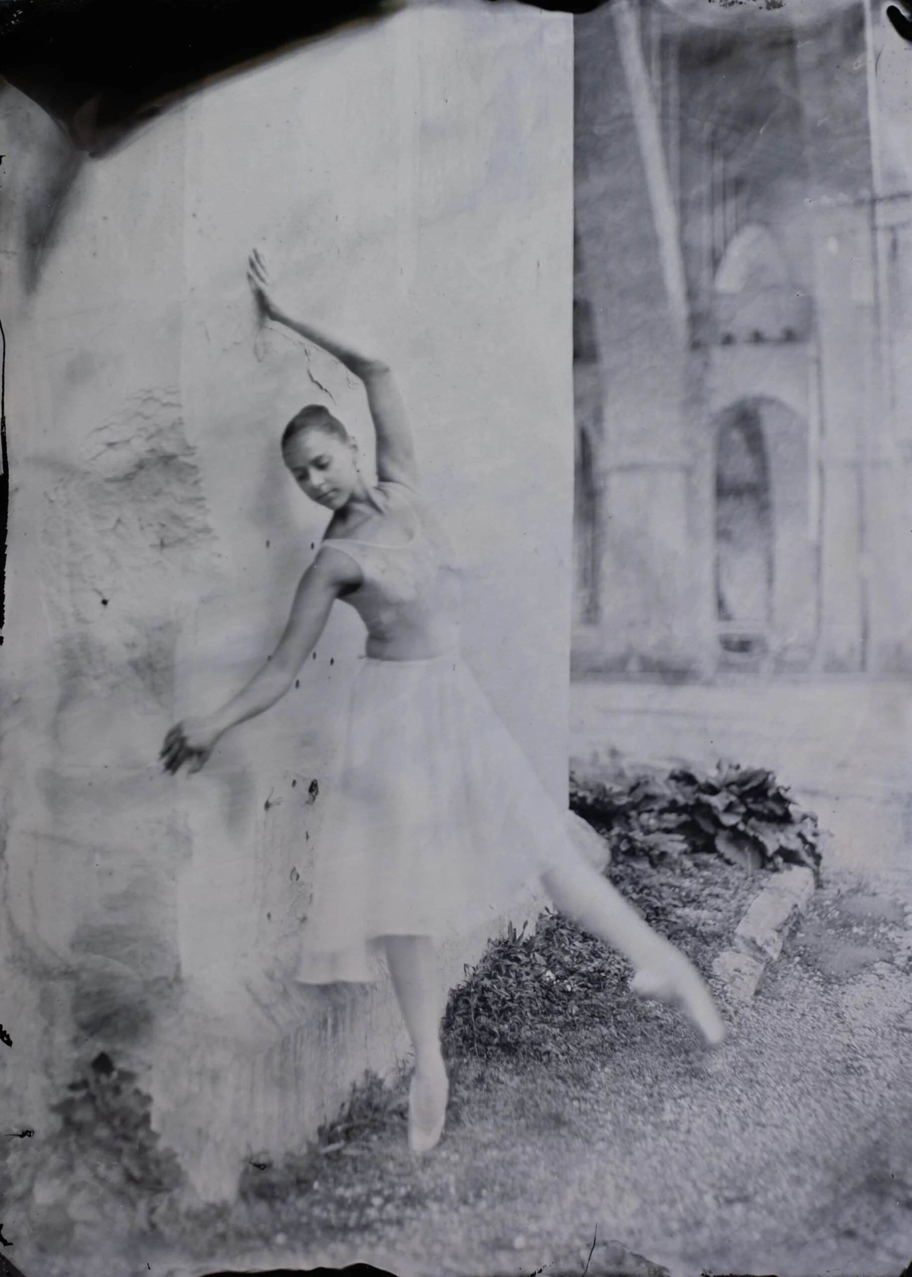 A ballerina dancing outdoors, wearing a ballet costume, with one arm raised above her head and the other extended to the side, with a building and trees in the background.