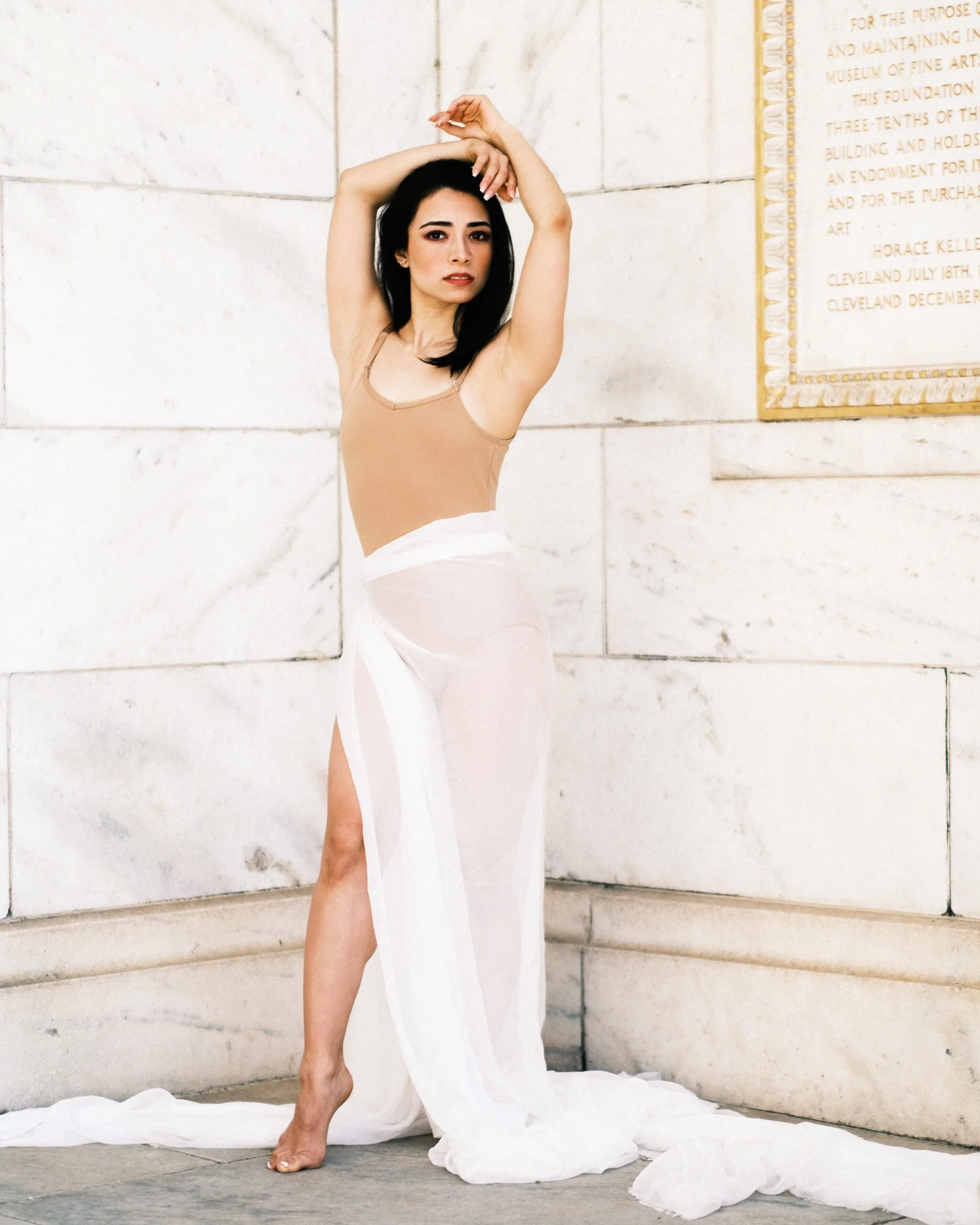 A woman with long dark hair in a dance pose stands against a white marble wall. She has one arm raised above her head and the other bent, touching her forehead. She wears a beige top and a white skirt or wrap that drapes to the ground. A textured mar