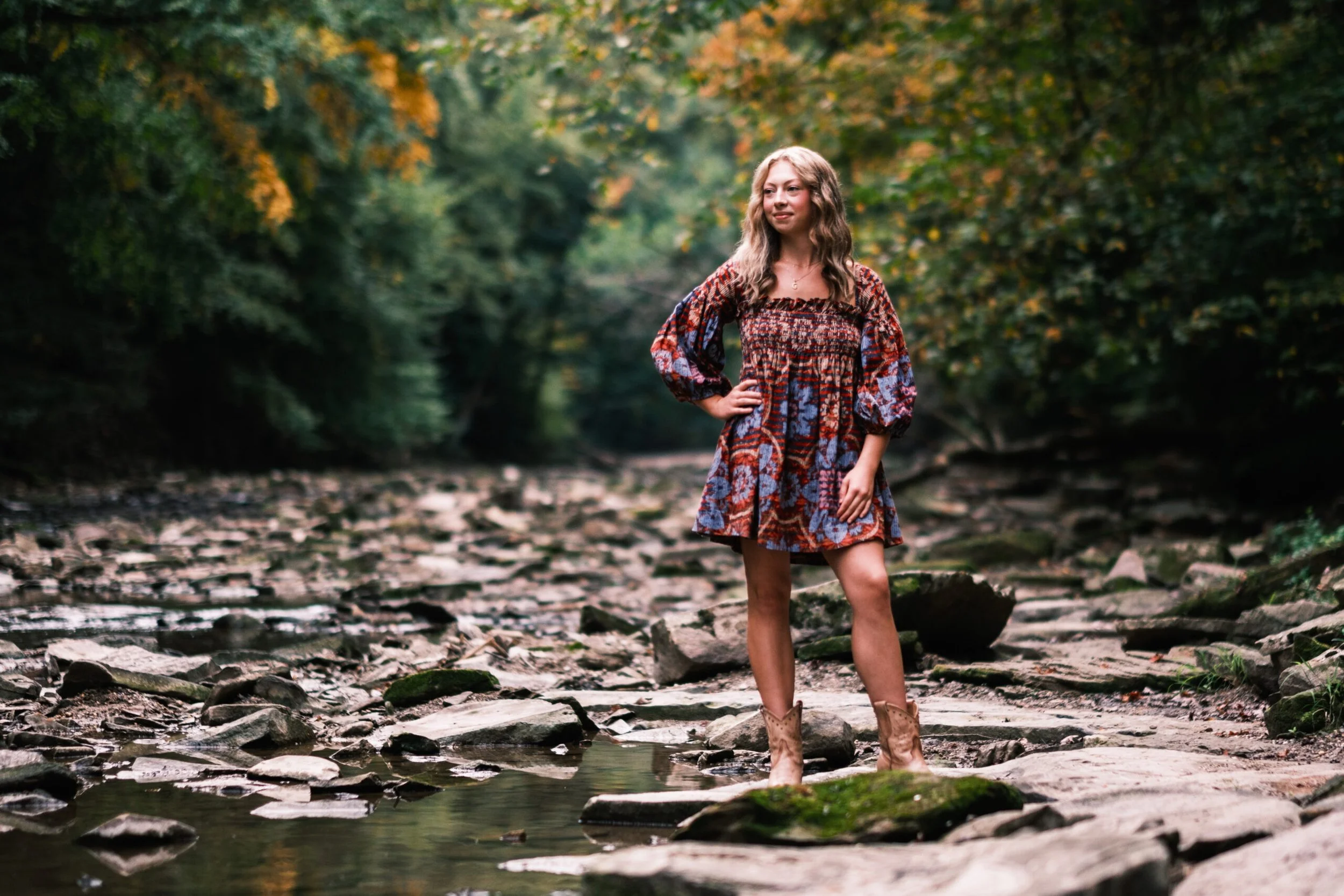 A young woman with wavy blonde hair standing in a shallow creek surrounded by autumn foliage, wearing a colorful patterned dress and cowboy boots.