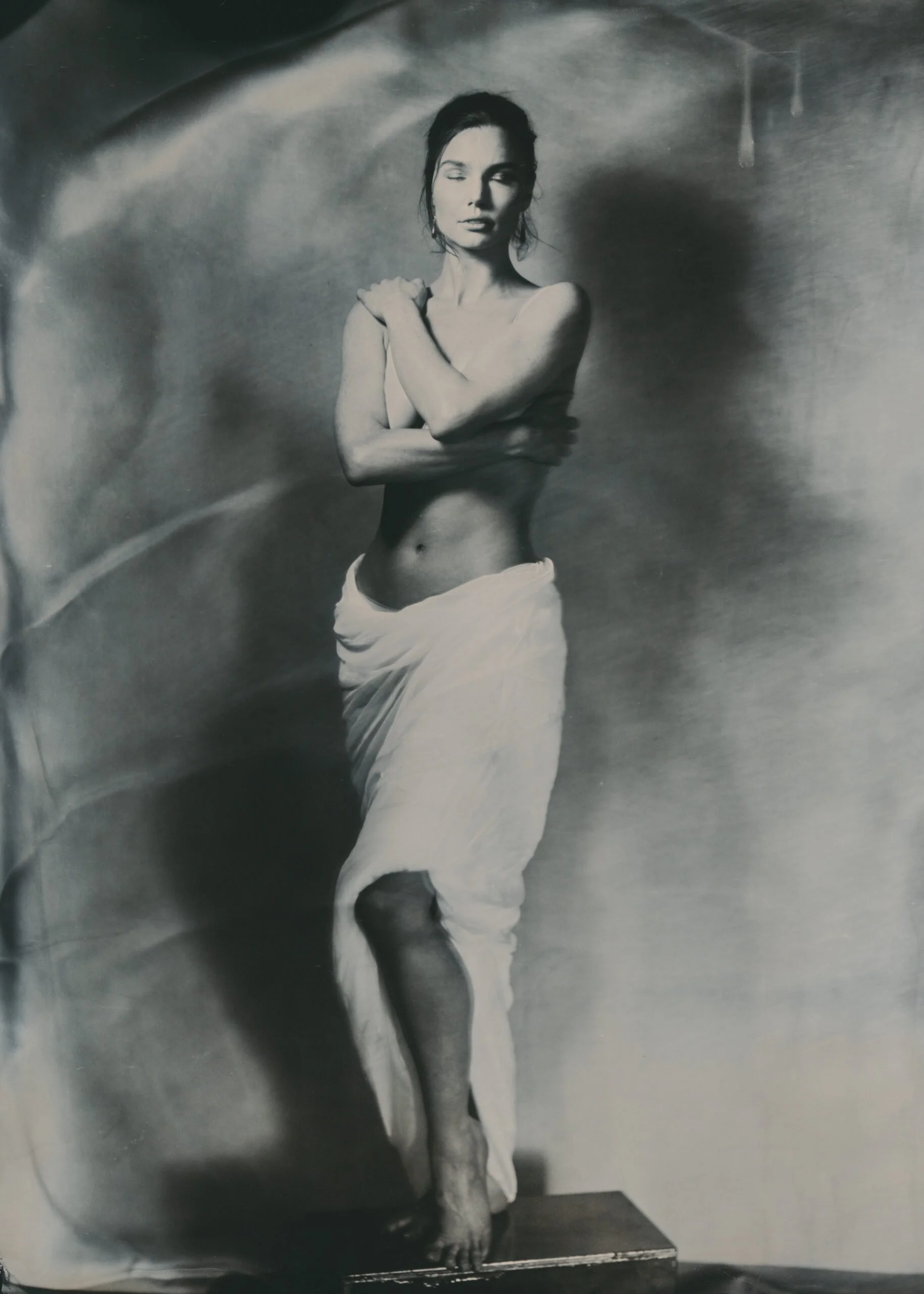 Black and white photograph of a woman standing on a platform, with her arms crossed over her chest, wearing a draped skirt or towel around her waist and a strapless top, with her eyes closed and head tilted slightly to the side.
