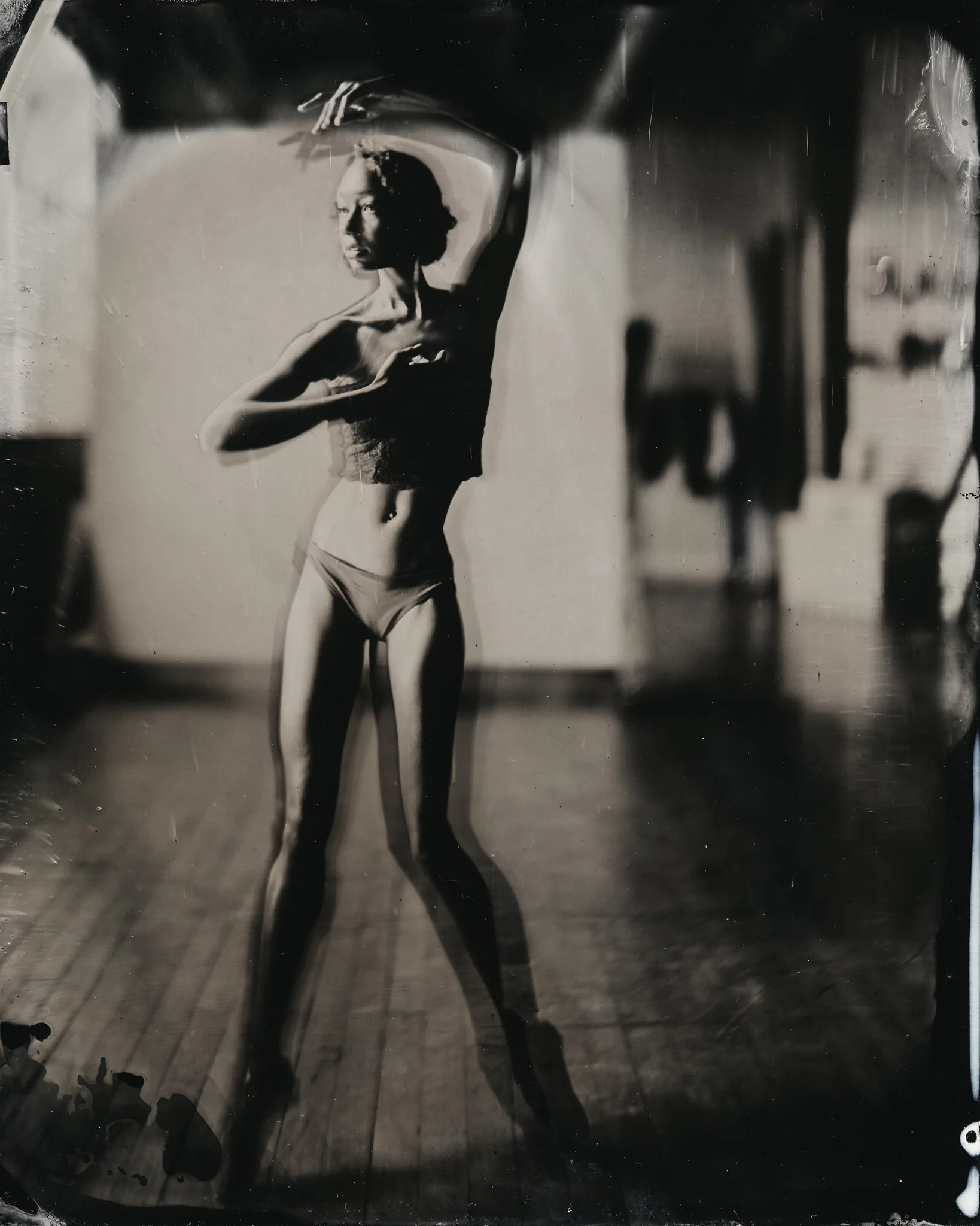 A mirror reflection of a woman taking a selfie, wearing a black top and shorts, with dark hair, in a room with wooden floors and blurred background.