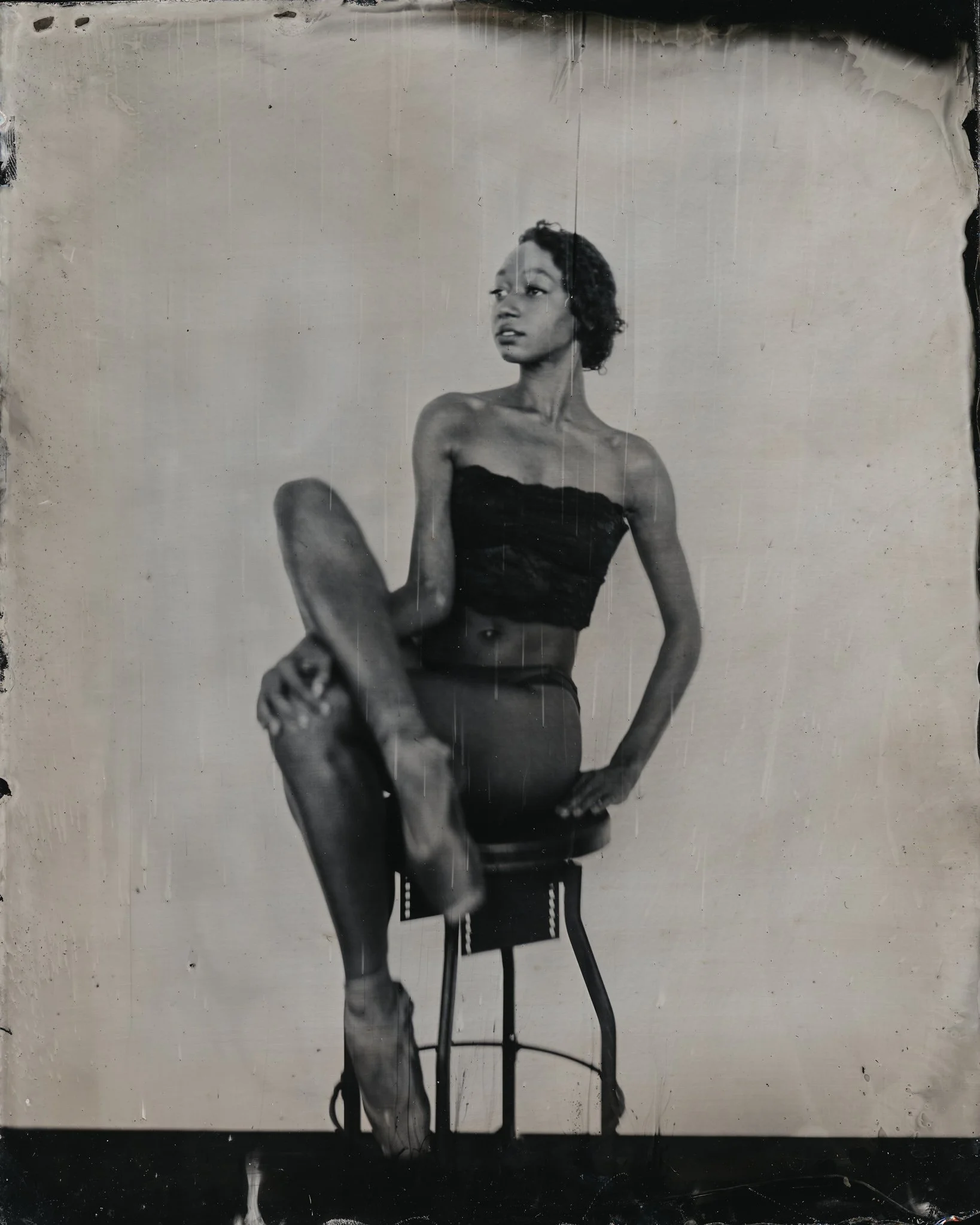 Black-and-white photograph of a woman sitting on a chair, with her legs crossed, in a poised pose, wearing a strapless top, with curly short hair, glasses, and natural makeup, against a plain background.