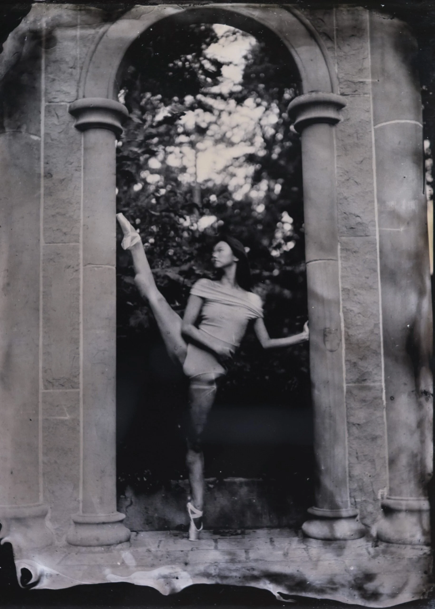 A woman with long dark hair wearing a short-sleeved top and shorts is standing in front of an archway with columns, holding a flower in her right hand. There are trees with leaves visible through the archway.