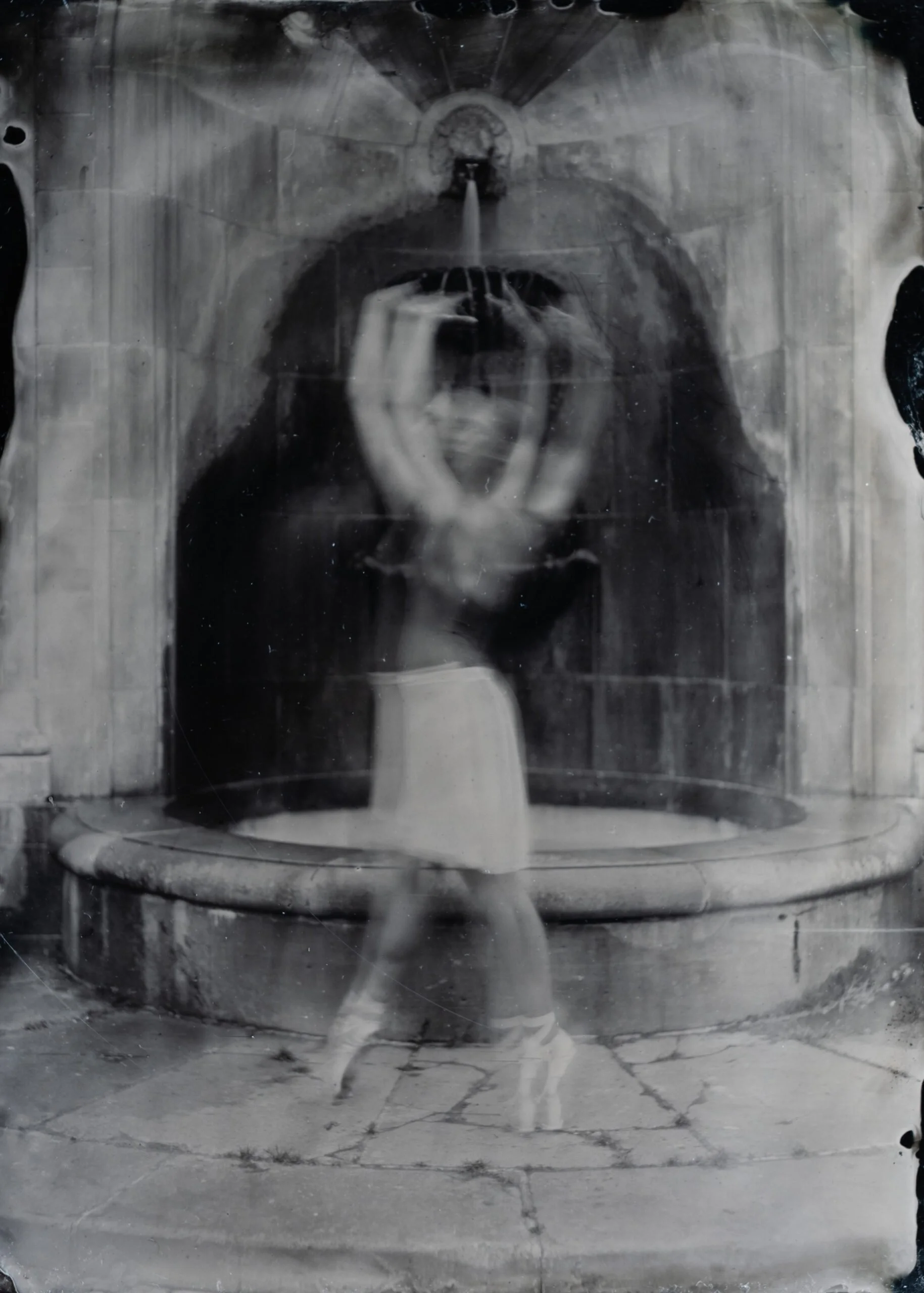 A black and white photograph of a person taking a photo of their reflection in a mirror. The person is standing with their arms raised, holding a smartphone, and the background scene is visible in the mirror's reflection.