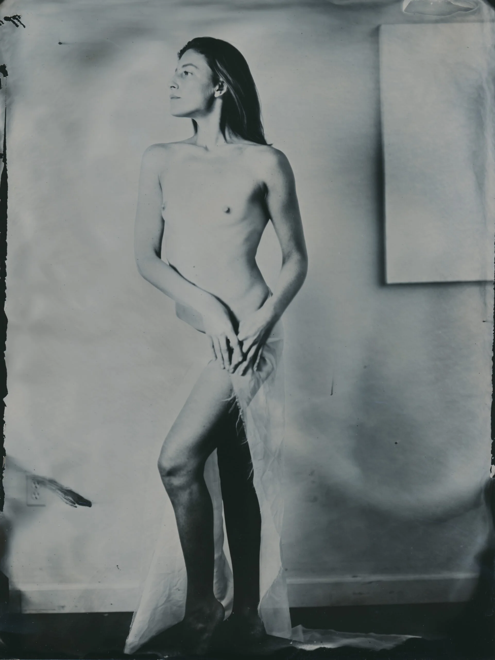 Black and white photograph of a topless woman with long hair, standing sideways with arms crossed, in a studio setting with a plain background.