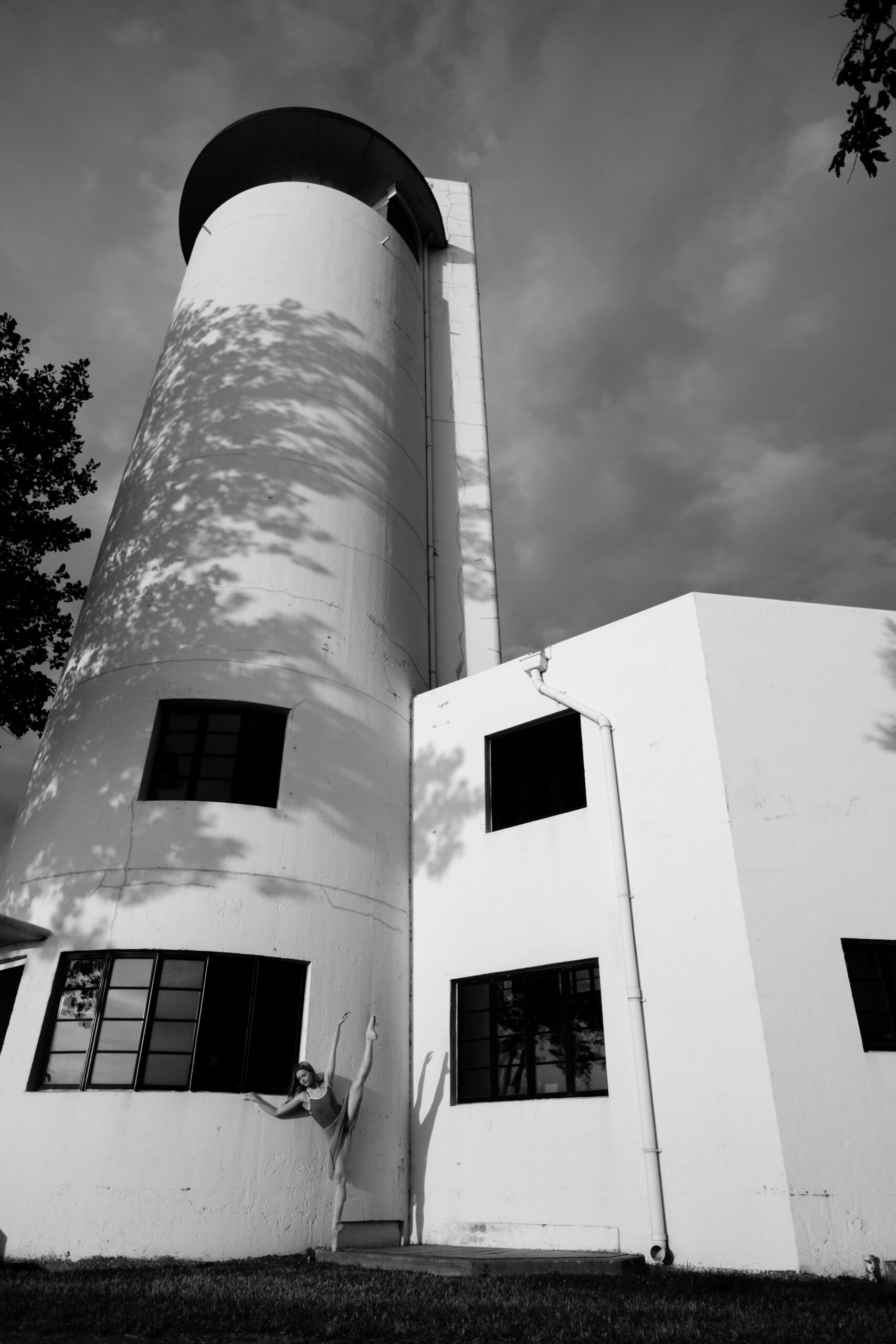 A woman dancing in a ballet pose outside a white building with a tall, rounded tower. The image is in black and white, with shadows from trees cast on the building.