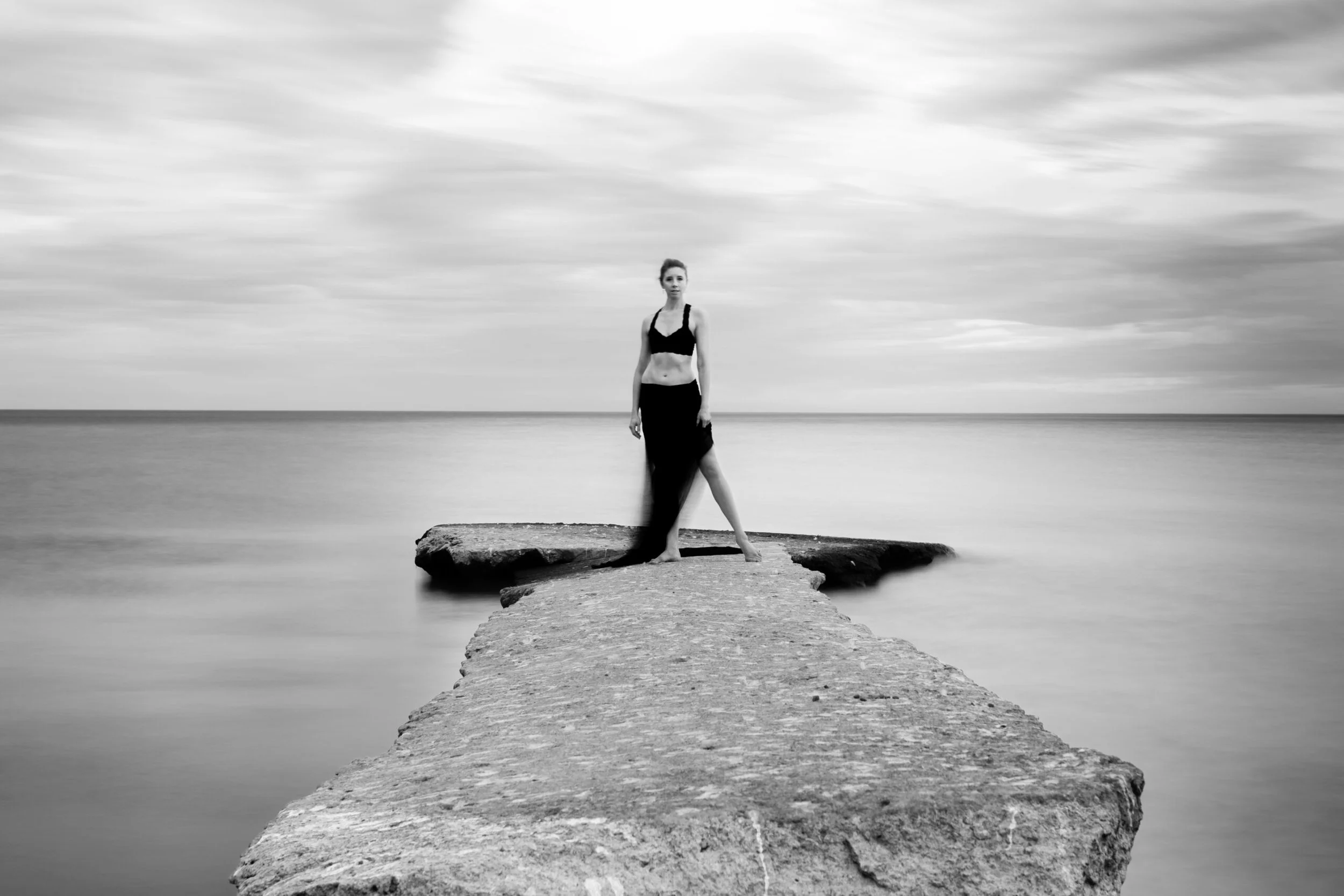 A woman in a black top and long skirt standing on a narrow concrete pier extending into a calm body of water, with a cloudy sky overhead in black and white.