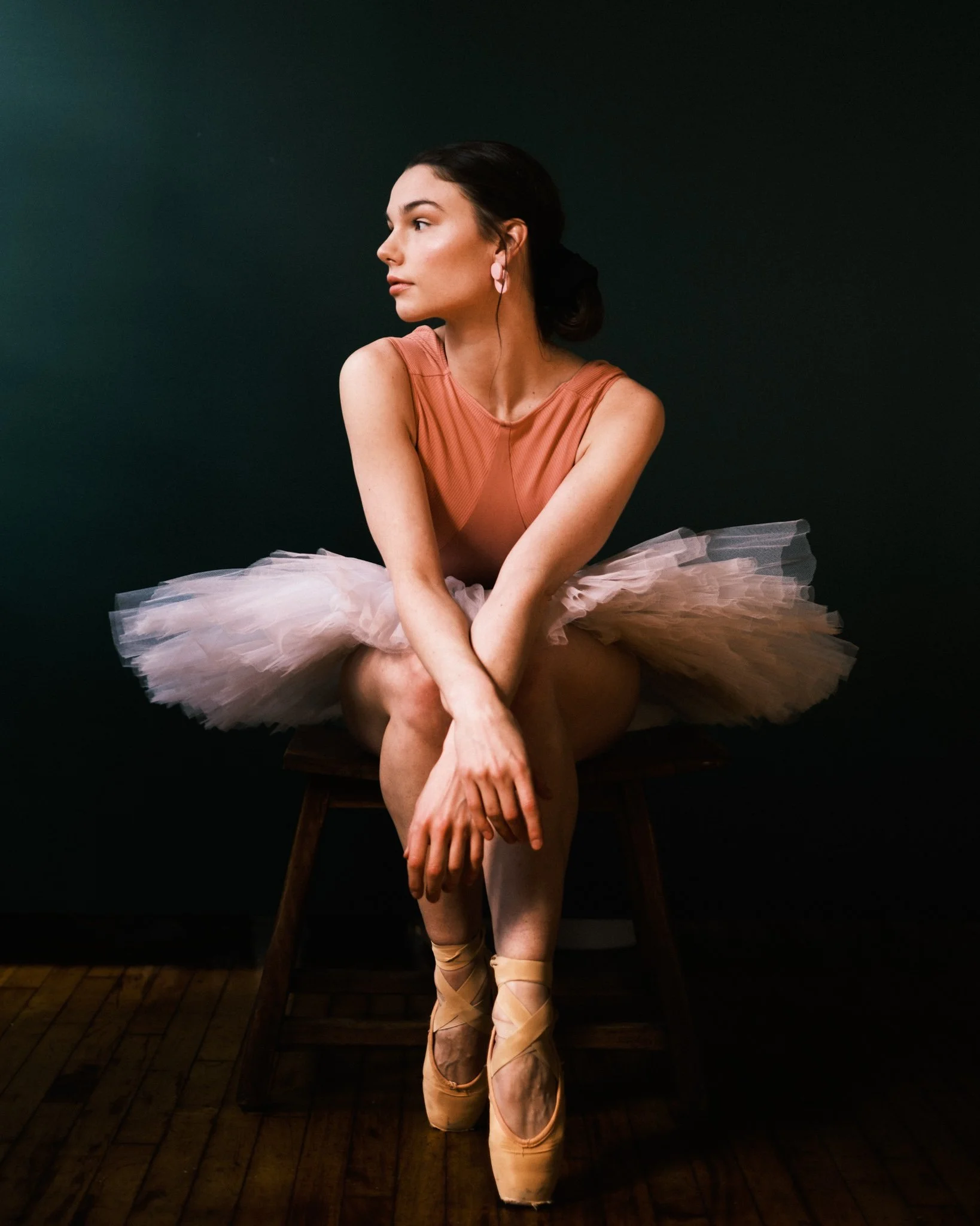 A ballerina sitting on a wooden stool, wearing a pink ballet tutu, pointe shoes, and an orange sleeveless top, with her legs crossed and arms resting on her lap, against a dark background.