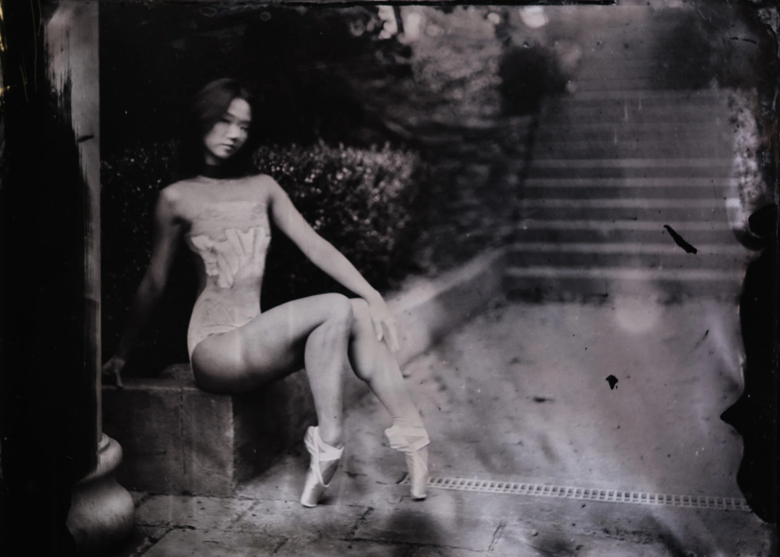 Black and white photo of a person sitting on a bench, wearing light-colored clothes, with legs crossed, in a park or outdoor area with trees, plants, and steps in the background.