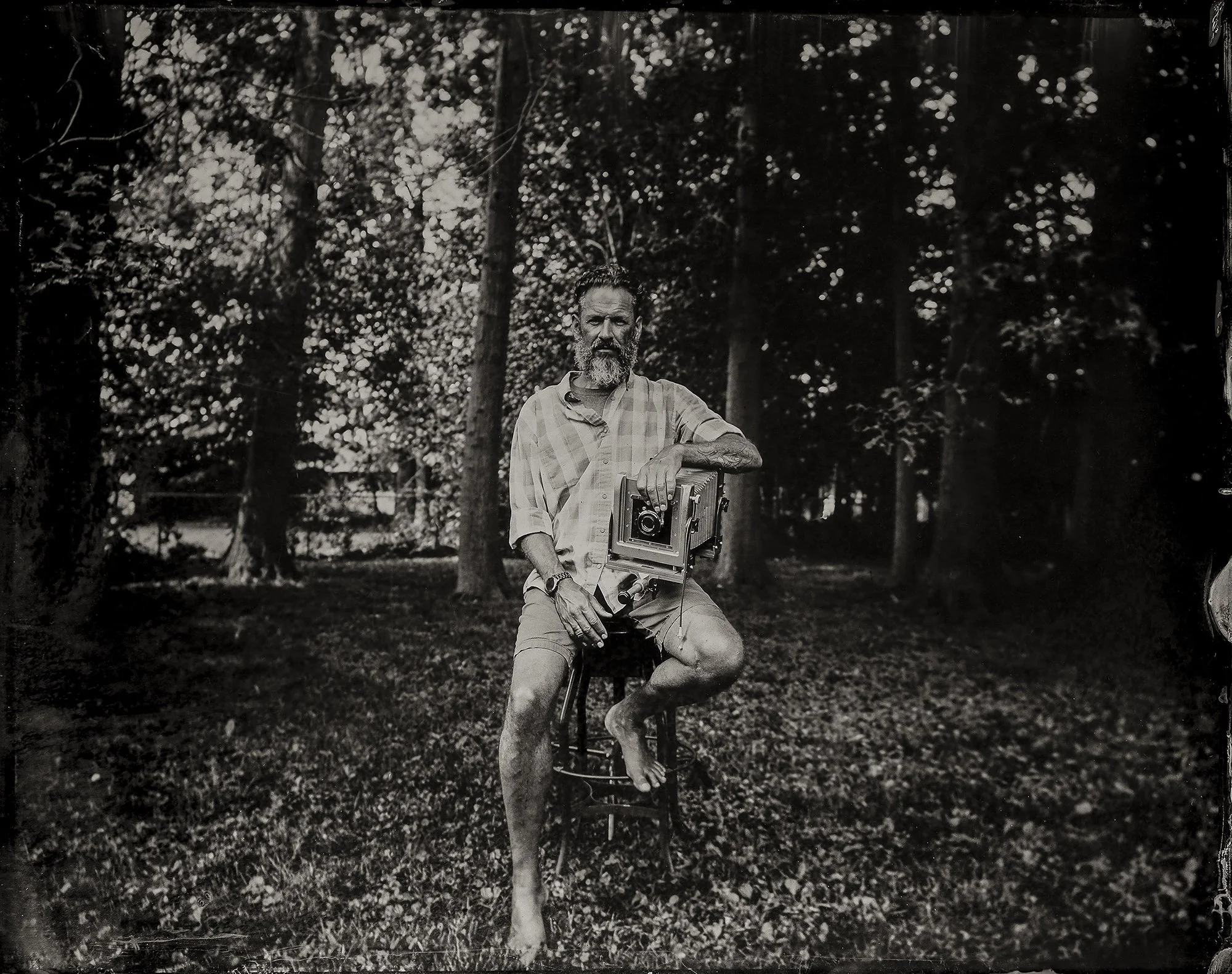 Black and white photo of a man with a beard, wearing a checked shirt, sitting outdoors in a park with trees in the background. He is holding a vintage camera and resting his arm on a tree trunk.