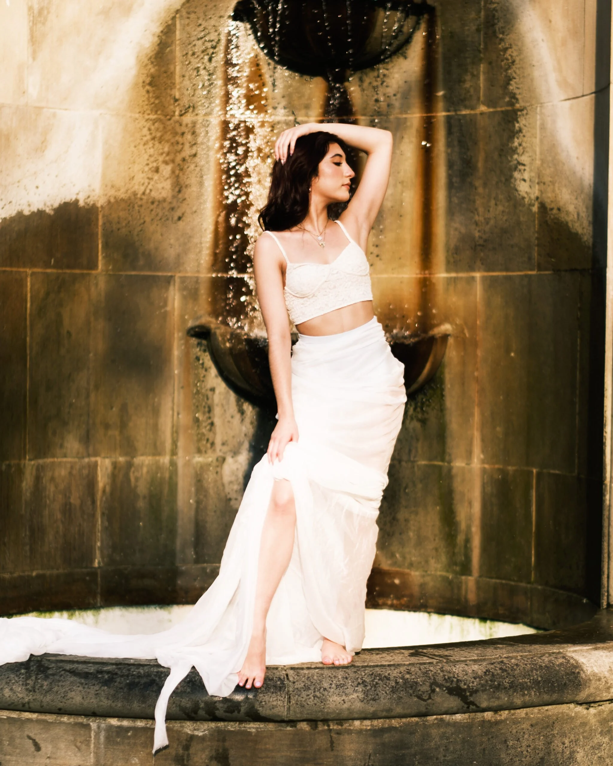 A woman in a white dress standing near a water feature with a fountain, her hand resting on her head, eyes closed, and a calm expression.