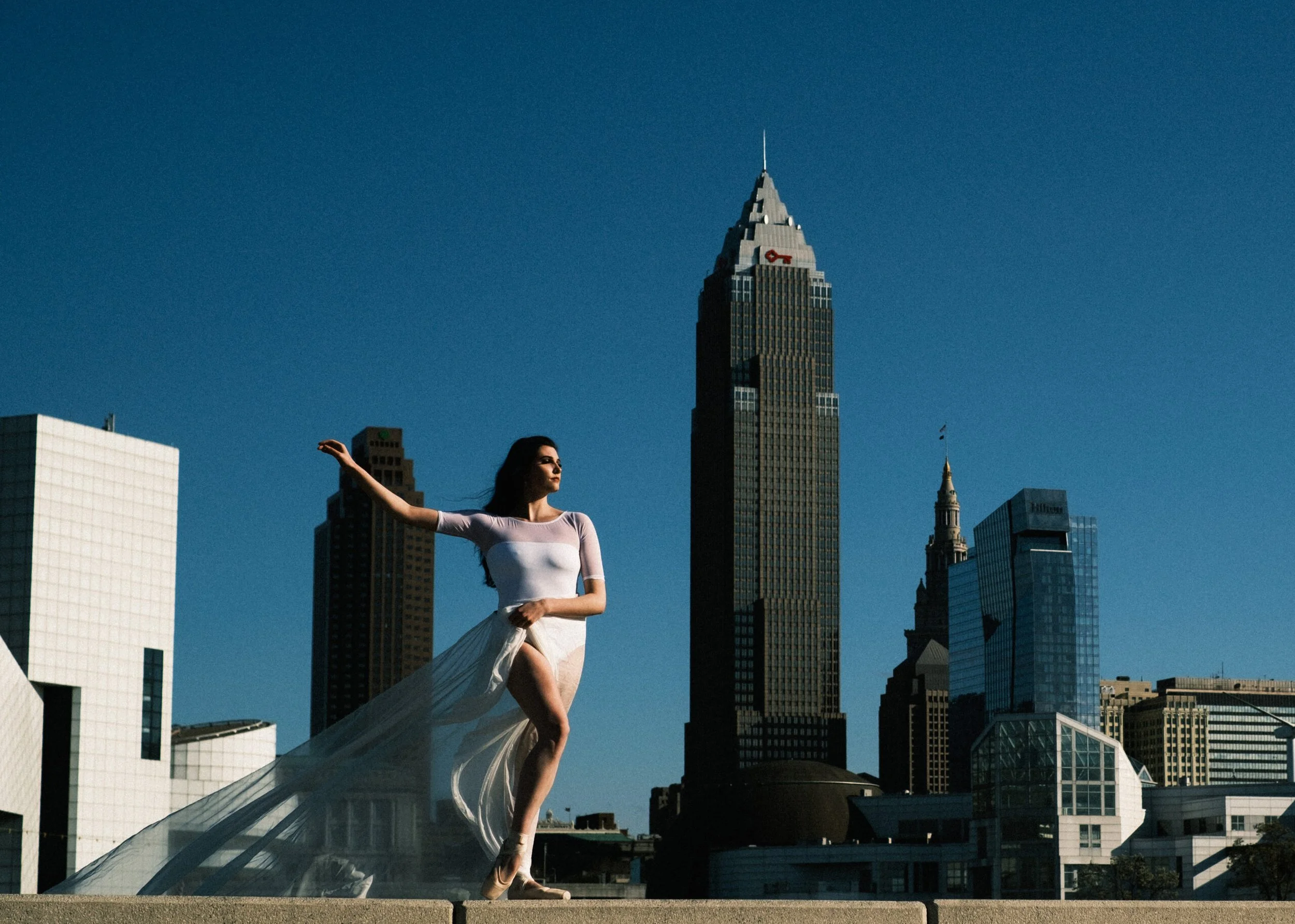 A dancer in a white top and flowing skirt performs on a ledge against a city skyline with tall buildings, including a prominent dark tower, under a clear blue sky.