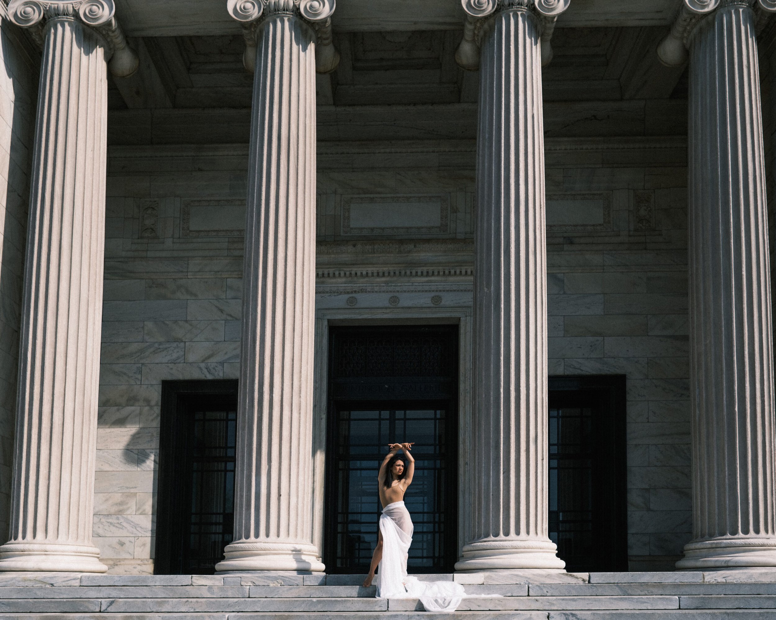 A woman in a white dress standing on the steps of a building with tall, classical columns, raising her arms above her head.