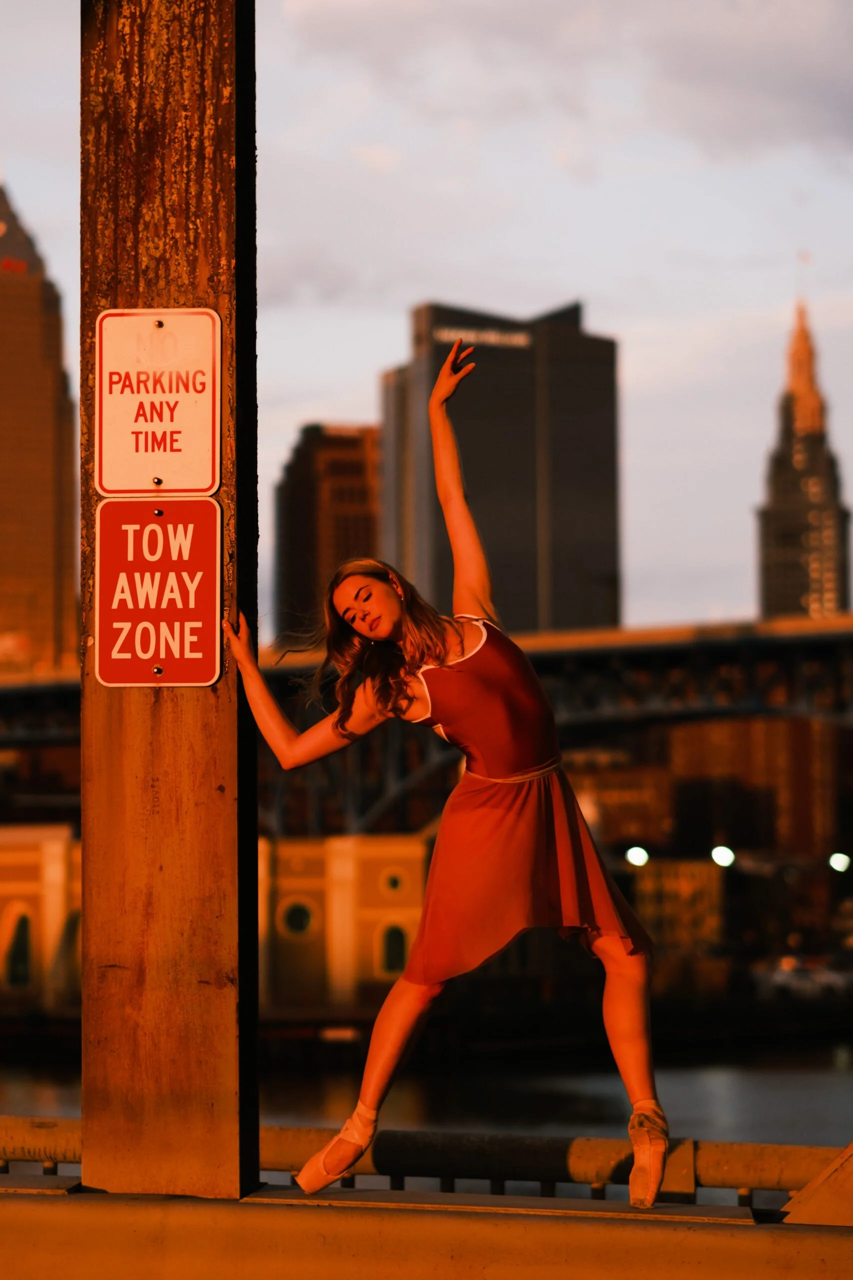 A woman in ballet attire standing on a city rooftop at sunset, with one arm raised above her head and leaning against a wooden post that has a 'Parking Any Time' and 'Tow Away Zone' sign, with city buildings and a cloudy sky in the background.