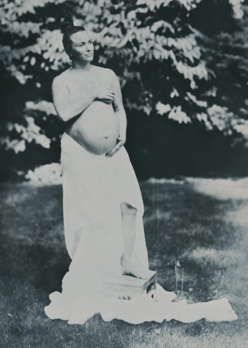 A pregnant woman standing outdoors near trees, draped in a white cloth, with one hand on her belly and the other on her chest, next to a wooden box on a cloth on the ground.
