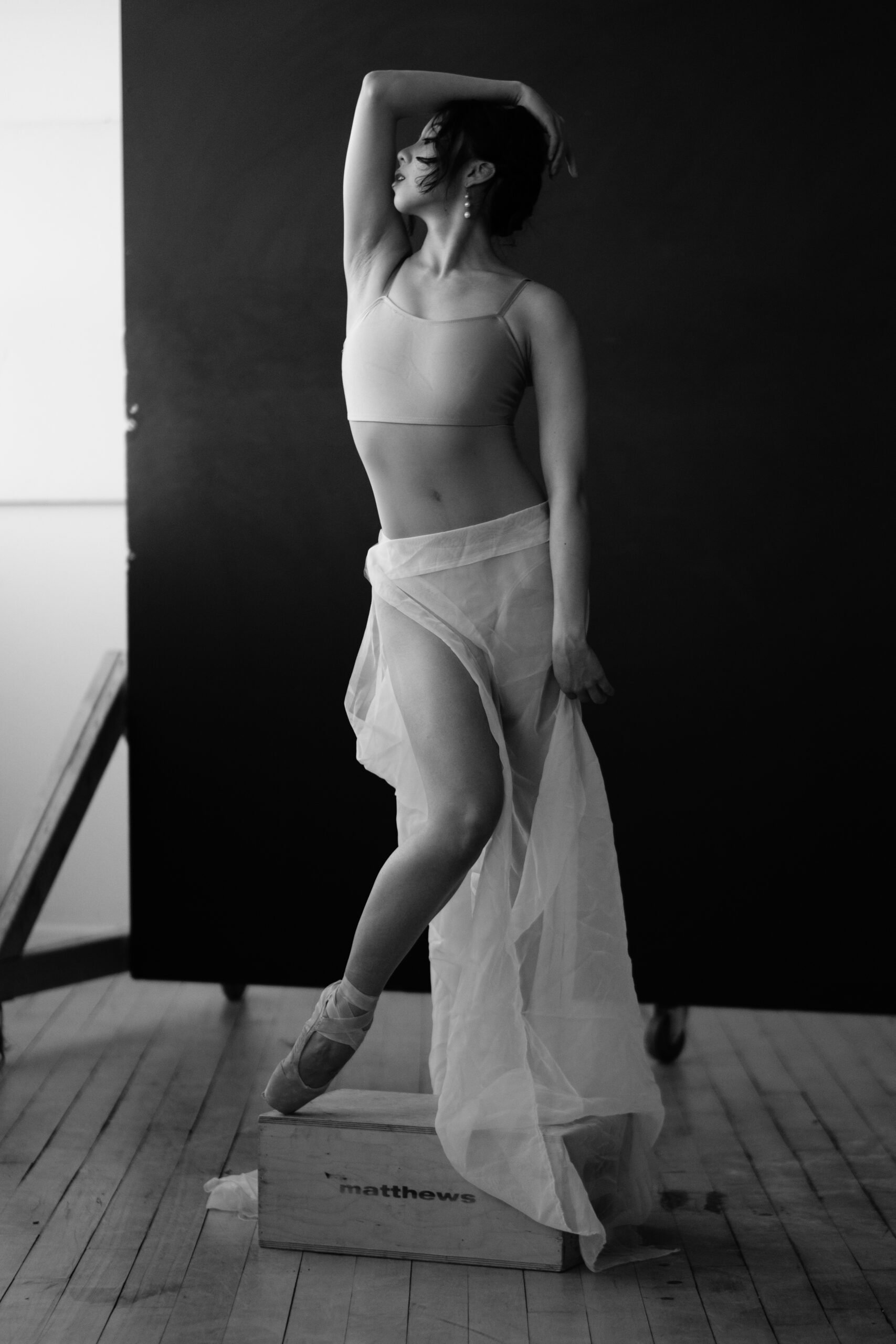 A black and white photo of a ballet dancer in pointe shoes, leaning against a wooden box with the word 'matthews' on it. She is wearing a tank top and a flowing sheer skirt, with her left arm bent and hand on her head, eyes closed, posing gracefully 