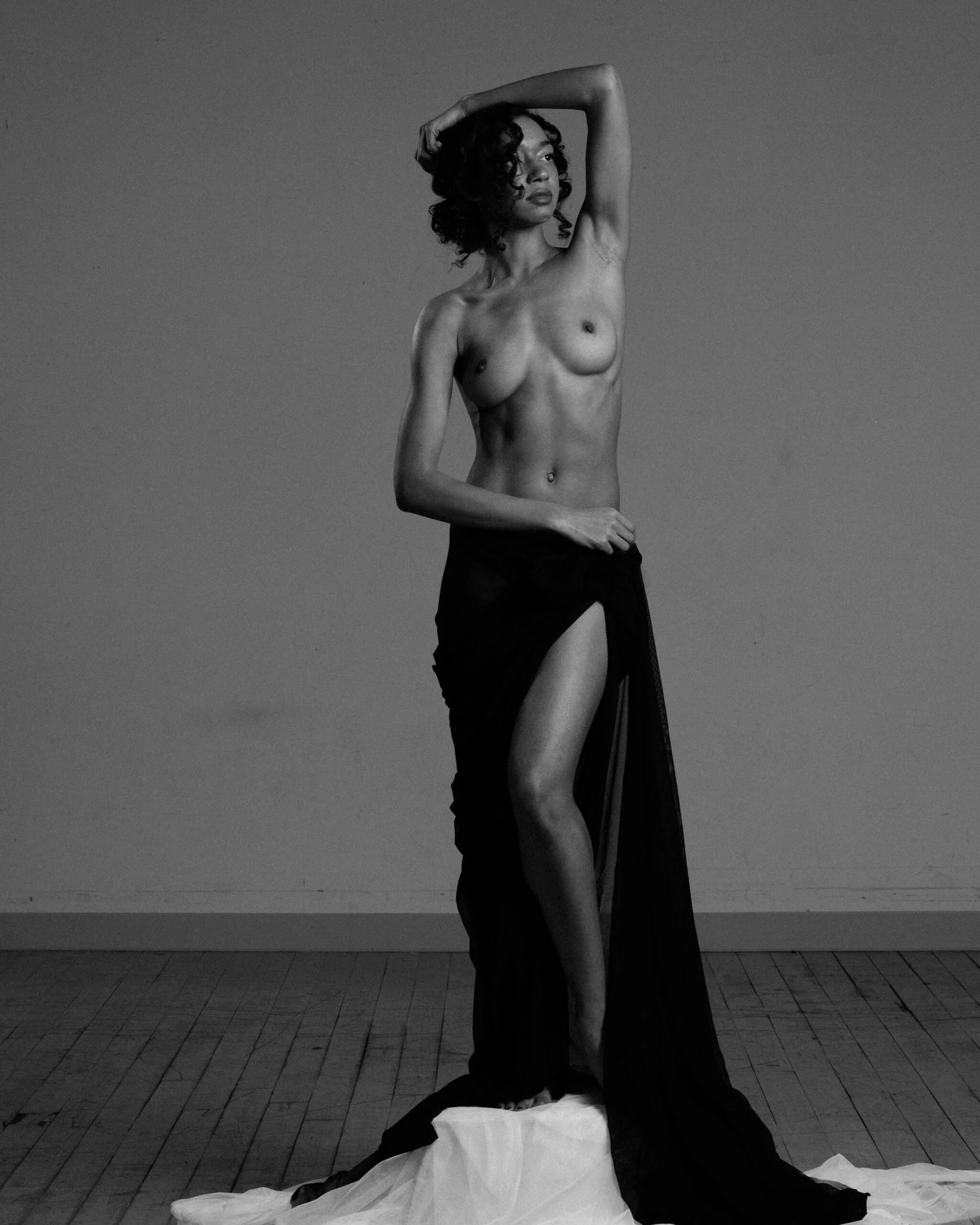 A woman standing topless on a platform covered with white fabric, posing with one arm raised above her head and wearing a long black skirt, against a plain background.