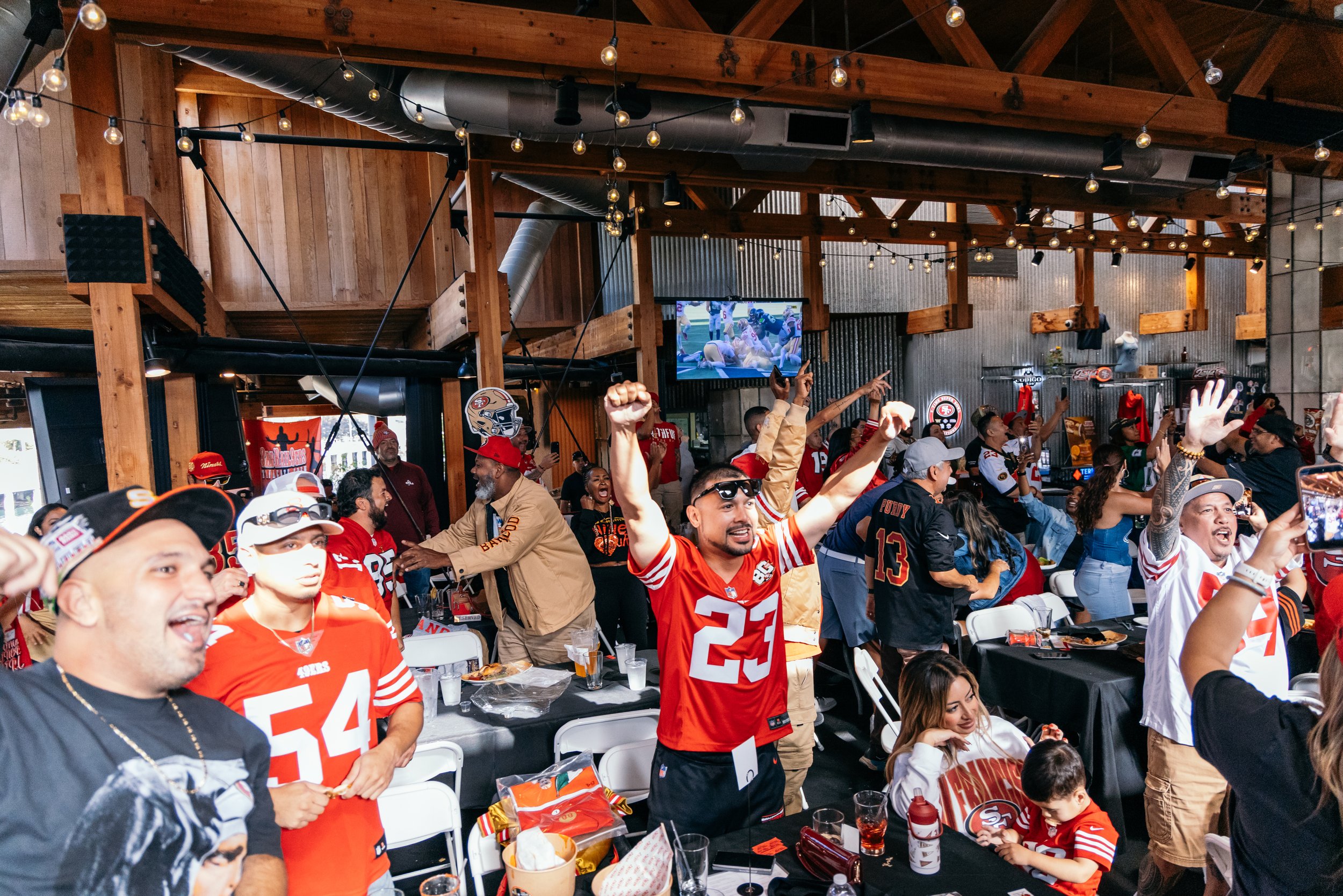 A lively indoor gathering of San Francisco 49ers fans wearing team jerseys and apparel, celebrating while watching a game on a large screen. The venue has a rustic wooden interior with string lights overhead.