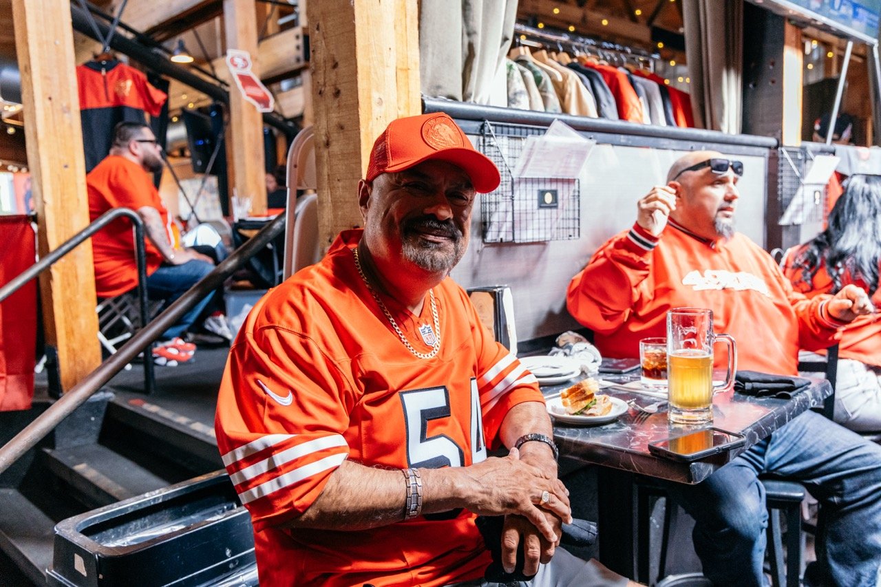 A man wearing an orange Denver Broncos jersey and cap sitting at a table in a sports bar, smiling and holding his hands together. There are drinks and food on the table, with more people in the background also wearing orange sports apparel.