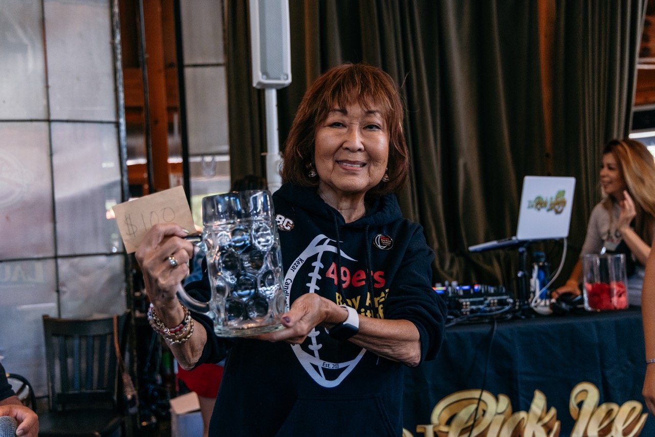 A smiling elderly woman holding a clear plastic tray with bottles and a cardboard sign that reads '$10'. She is at an indoor event with a DJ setup and another woman in the background.