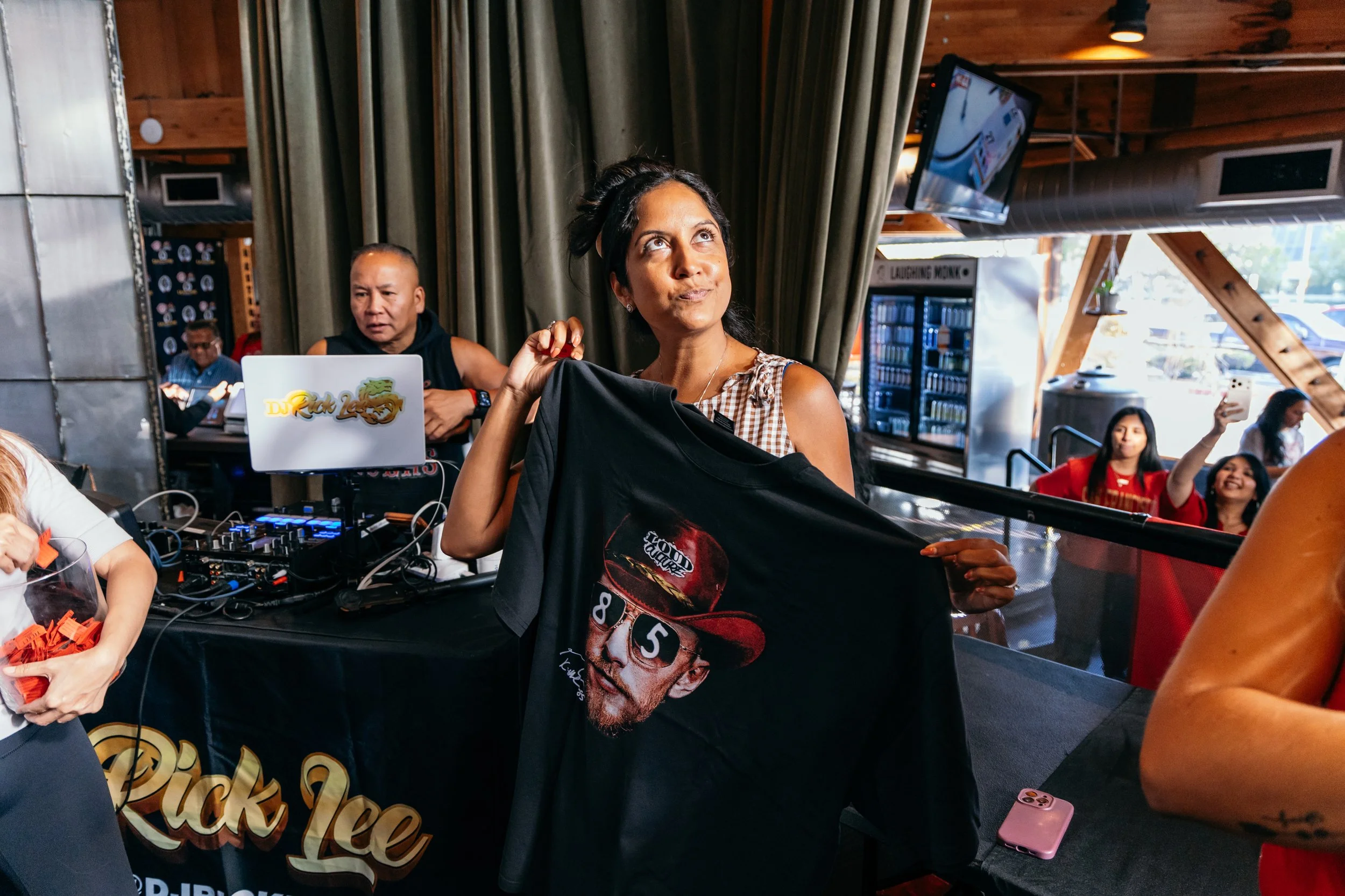 A woman holding a black T-shirt with a graphic of a person's face and sunglasses at a social event, surrounded by people and a DJ setup in a wooden interior.