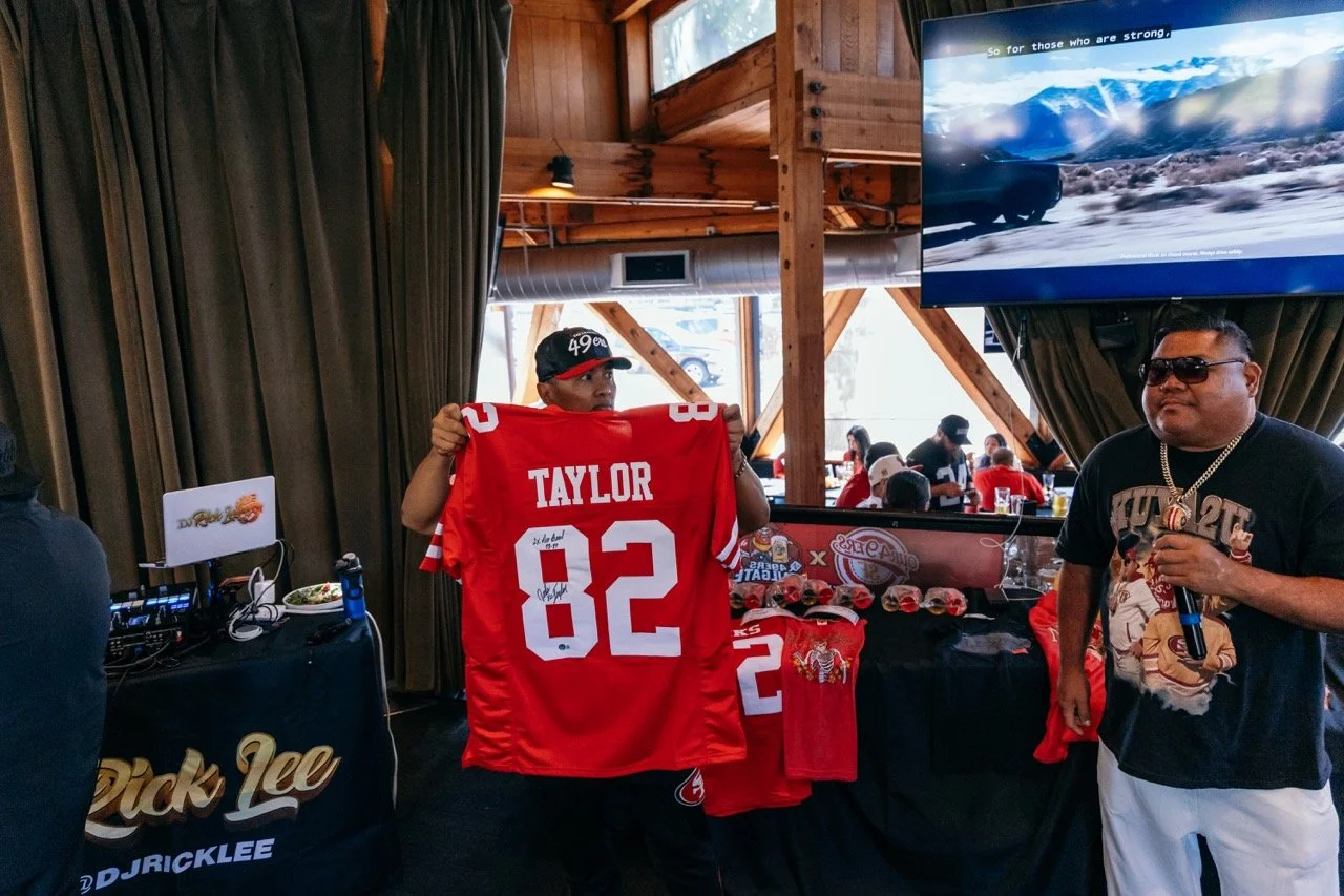 Person holding a red football jersey with 'TAYLOR' and the number 82, indoors at a gathering, with another man standing nearby, TV screen displaying a landscape, and tables with merchandise and snacks in the background.