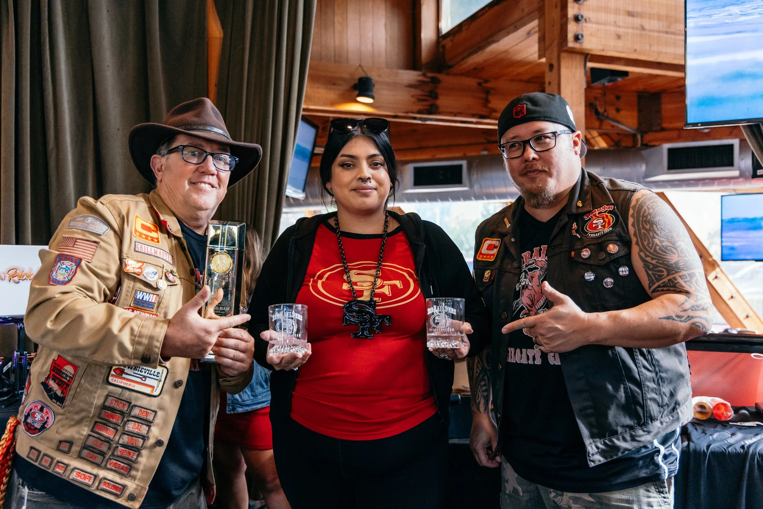 Three people posing for a photo indoors. The man on the left is wearing a tan jacket with patches and a brown cowboy hat, holding a glass of water. The woman in the middle is wearing a red shirt with a gold emblem, black necklace, and sunglasses on h