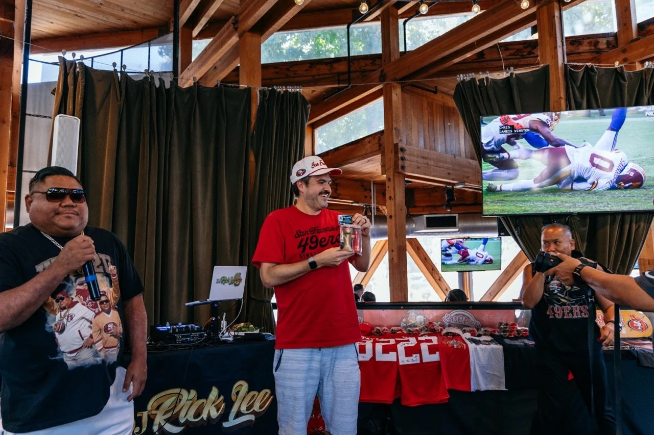 Group of San Francisco 49ers fans at a celebration event, with one man holding a microphone and wearing a black 49ers shirt, another man in a red 49ers shirt holding a gift, and another man taking a photo. The event is indoors with wooden walls, TV s