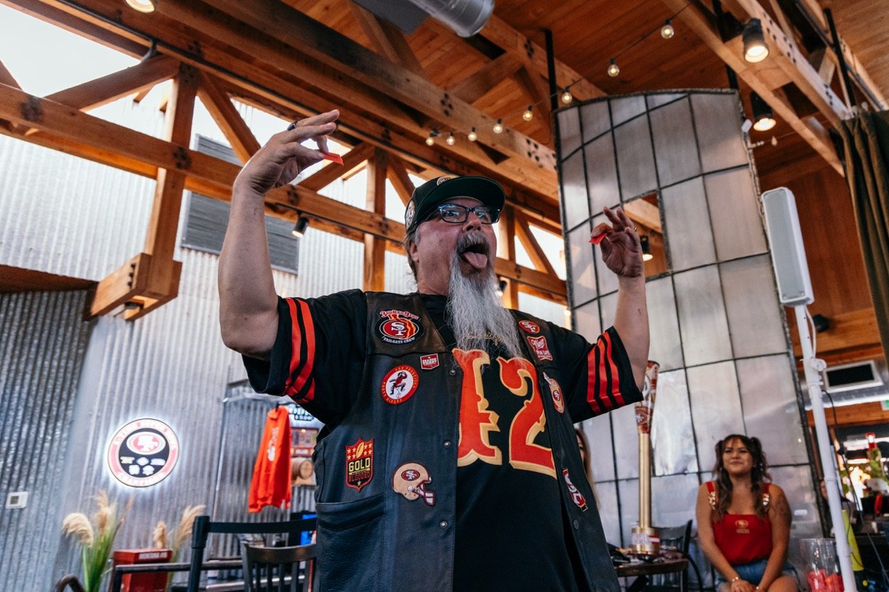 A man with a beard and glasses wearing a San Francisco 49ers jersey and jacket, celebrating enthusiastically with his tongue out and hands raised, inside a wooden-paneled restaurant or sports bar. A woman in a red top sits at a table in the backgroun