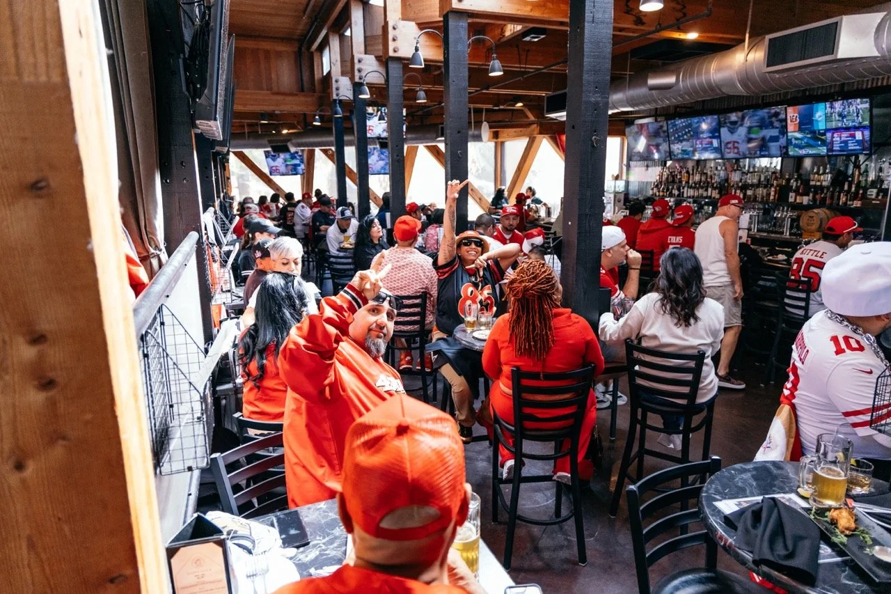 Crowd of sports fans wearing red and white jerseys in a bar watching a game on multiple large screens, with some making peace signs and others enjoying drinks.