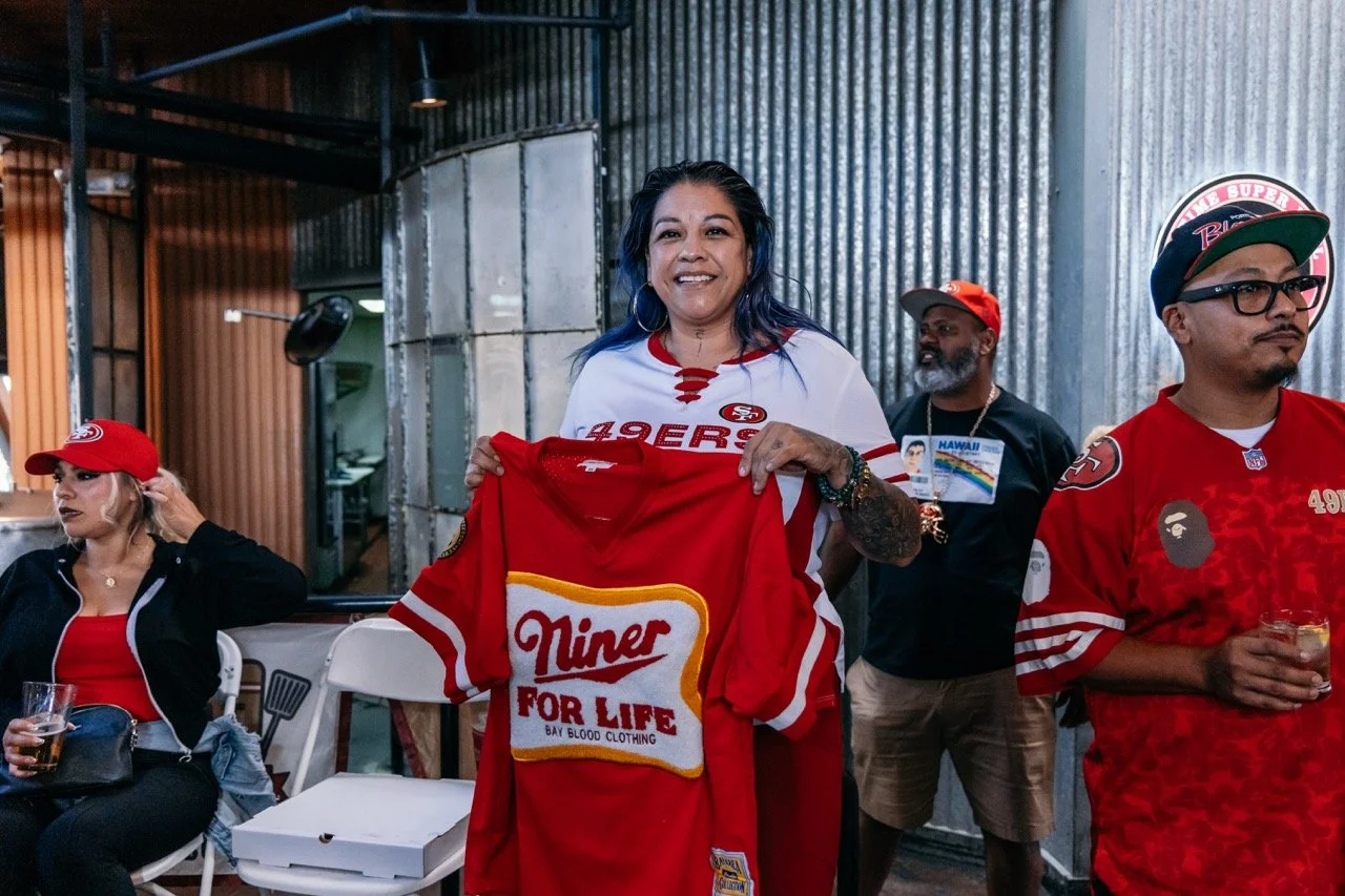 Group of people celebrating, one woman holding a red Kansas City 49ers jersey with a 'Niner for Life' logo, wearing a San Francisco 49ers shirt, in a rustic indoor setting, some wearing team apparel and glasses, with drinks in hand.