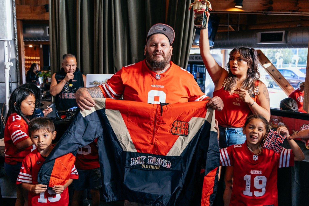 A man in a San Francisco 49ers football jersey and a baseball cap holding a jacket, surrounded by children and a woman, all in 49ers apparel, celebrating inside a restaurant or bar.