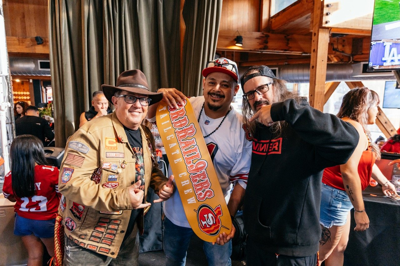 Three men celebrating together at a gathering with San Francisco 49ers and San Francisco Giants gear, holding a sign that reads 'Broncos Nation' with a logo of a bull.