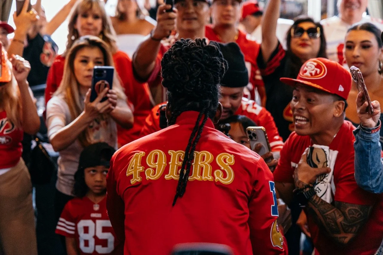 A group of San Francisco 49ers football fans wearing team apparel, gathered around a player during a meet-and-greet, with fans taking photos and videos.