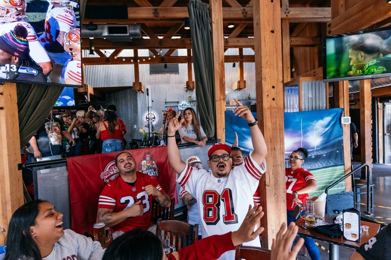 A group of sports fans, mostly wearing San Francisco 49ers jerseys, celebrating and enjoying a game at a bar or restaurant. Some are cheering, raising their hands, and laughing, with TV screens displaying football games.