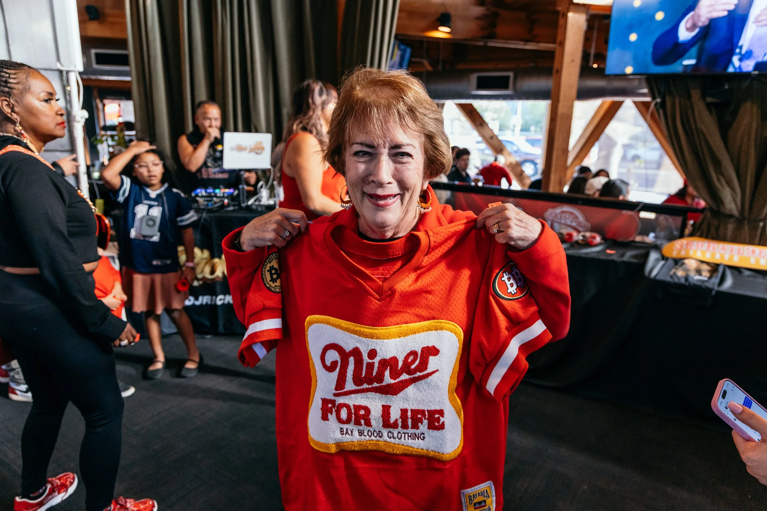 A woman in a red shirt with a 'Mothers for Life' patch on it, smiling and adjusting her shirt in a crowded indoor setting.