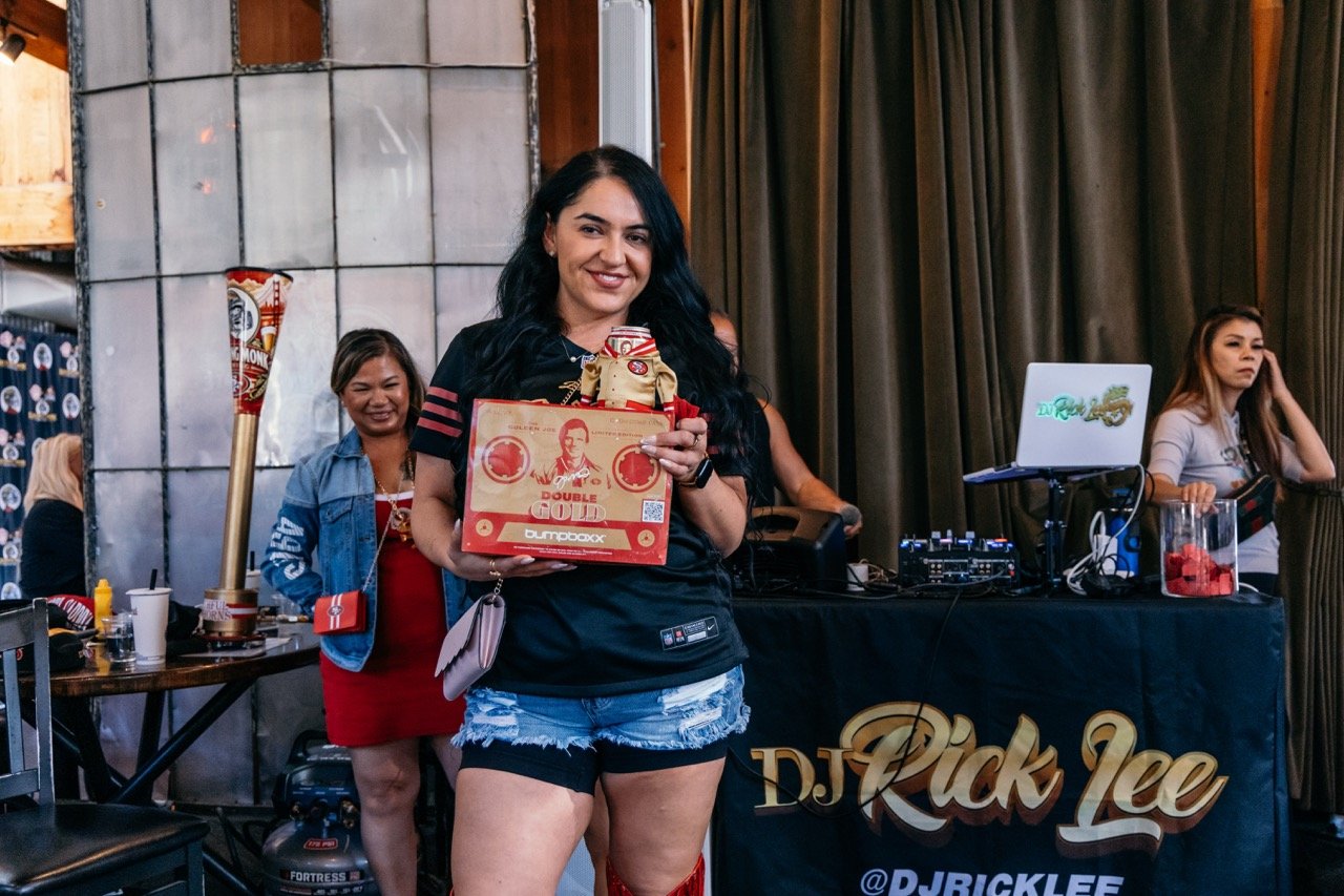 A woman with black hair holding a gift and a box of Double Gold coffee while smiling at an event, with DJ equipment and a woman in a gray shirt in the background.