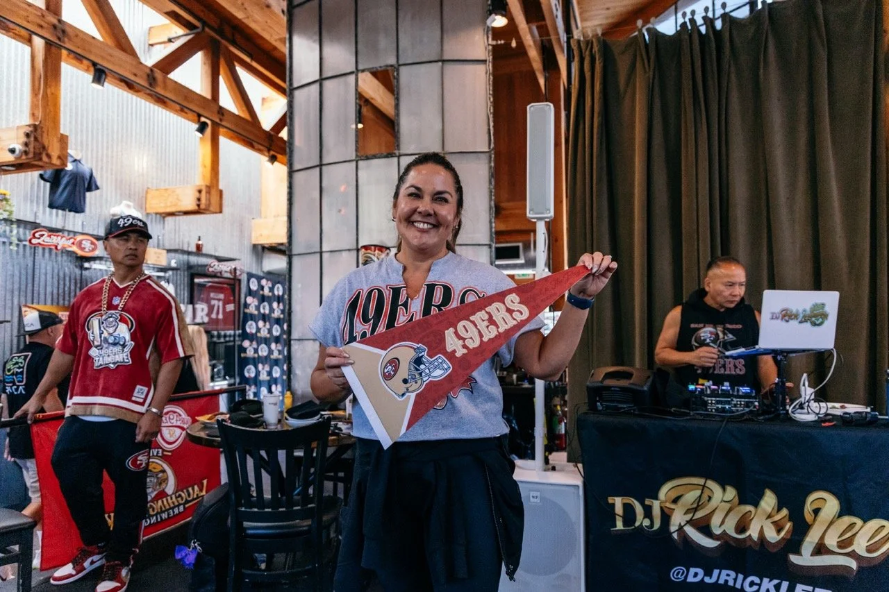 A woman smiling and holding a San Francisco 49ers pennant in a room decorated with 49ers memorabilia, with a DJ setup and a man wearing a 49ers shirt in the background.