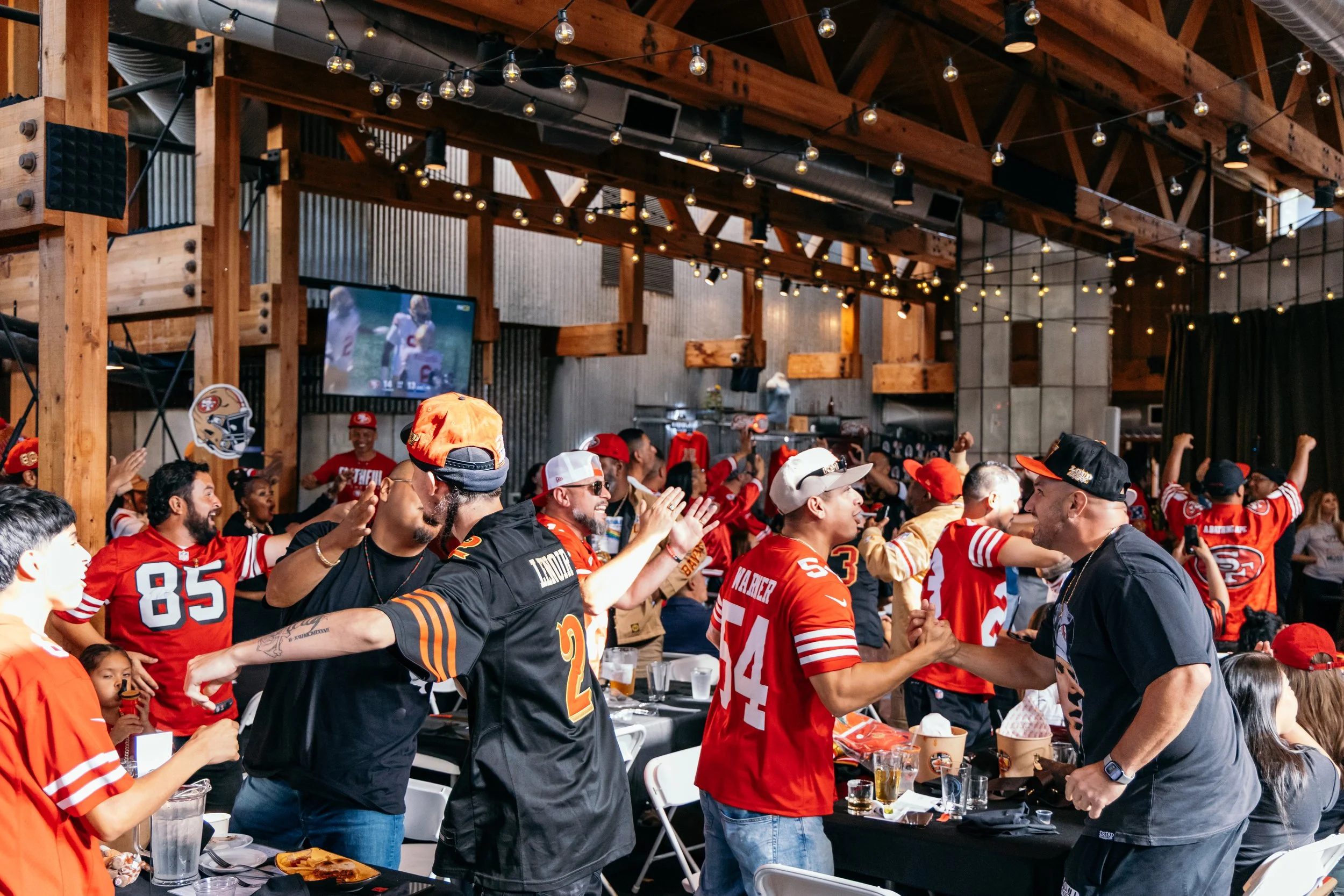 A lively indoor gathering of football fans wearing red and black jerseys, celebrating and interacting under string lights in a wooden, rustic-style venue.